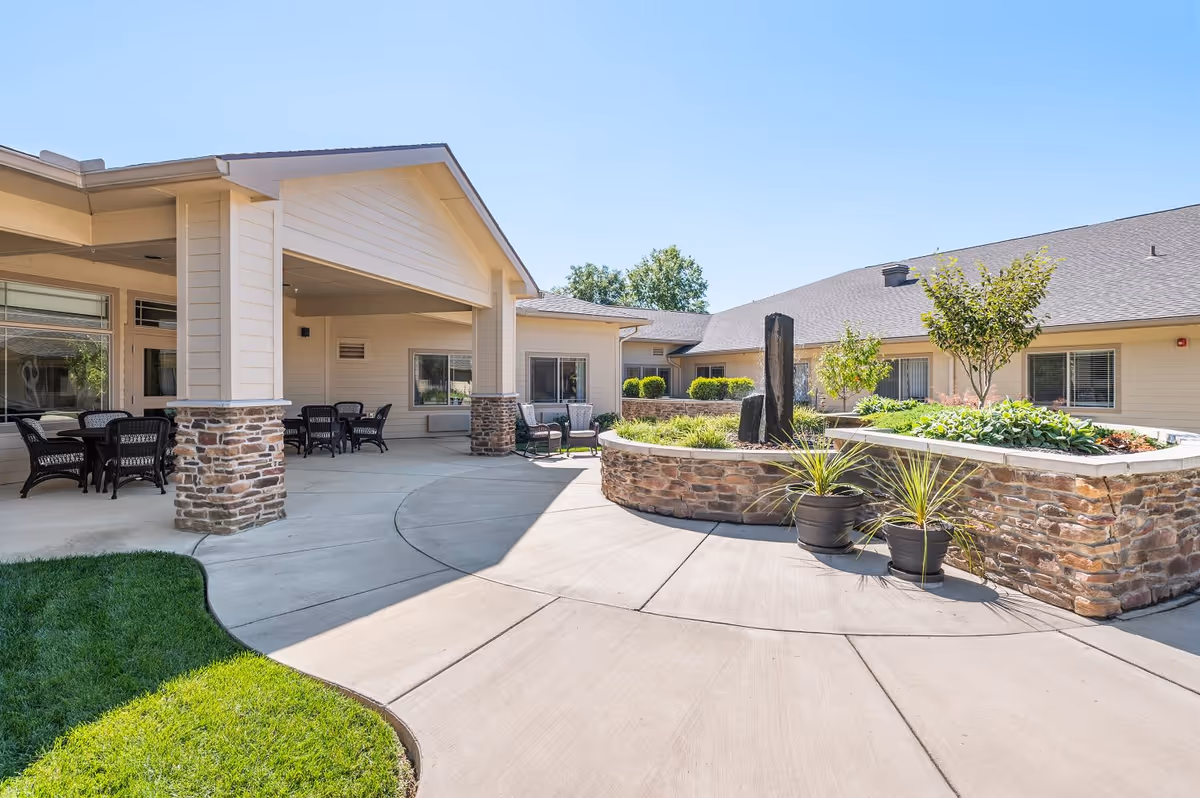 Outdoor courtyard area of Sage Park Assisted Living & Memory Care with a covered patio featuring seating arrangements, stone pillars, a raised stone planter with greenery and small trees, and a clear blue sky.