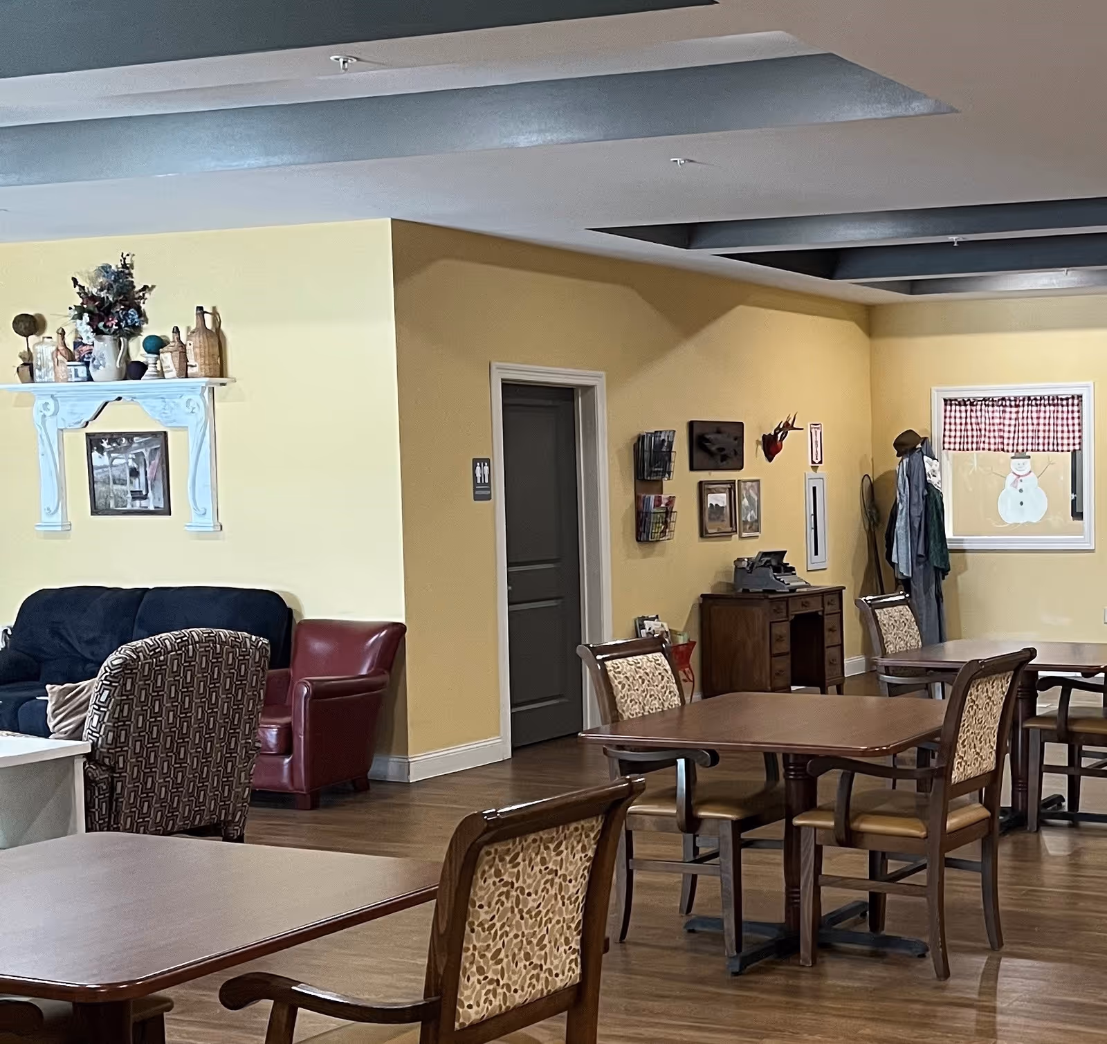 Interior of a common area in a retirement village with wooden tables and chairs arranged for seating. There is a dark blue couch, a red armchair, and a patterned armchair near a yellow wall. The wall has decorative items including a shelf with vases and a framed picture. A door with a restroom sign is visible, along with a small desk, coat rack, and a window with a snowman decoration.