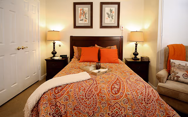 A cozy bedroom with a bed covered in an orange paisley patterned bedspread, two orange pillows, and a white throw blanket. There are two dark wooden nightstands on either side of the bed, each with a lamp. Above the bed are two framed botanical prints. To the right, there is a beige armchair with an orange throw and a decorative pillow.