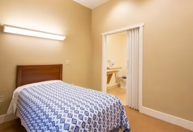 A small bedroom with a single bed covered with a blue and white patterned blanket. The room has beige walls and a wall-mounted light fixture above the bed. There is an open doorway with a white folding door leading to a bathroom with a toilet and sink visible.