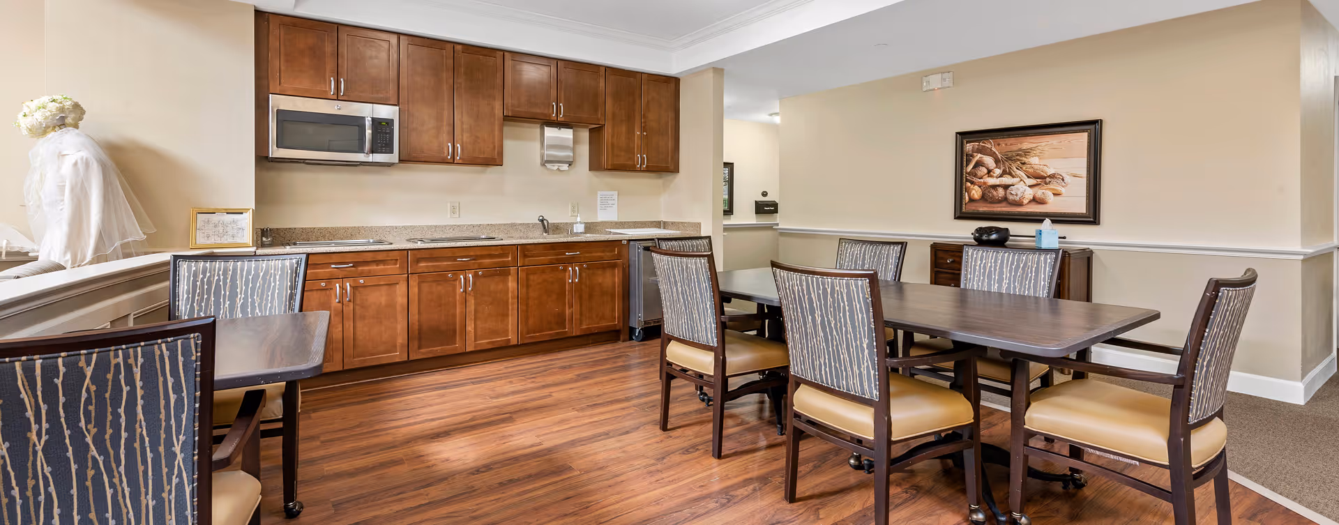 A dining area with a wooden table surrounded by six chairs with patterned backs and tan seats. Behind the table is a kitchenette with wooden cabinets, a microwave, a sink, and a granite countertop. The room has wood flooring and beige walls with a framed picture of bread on the wall.