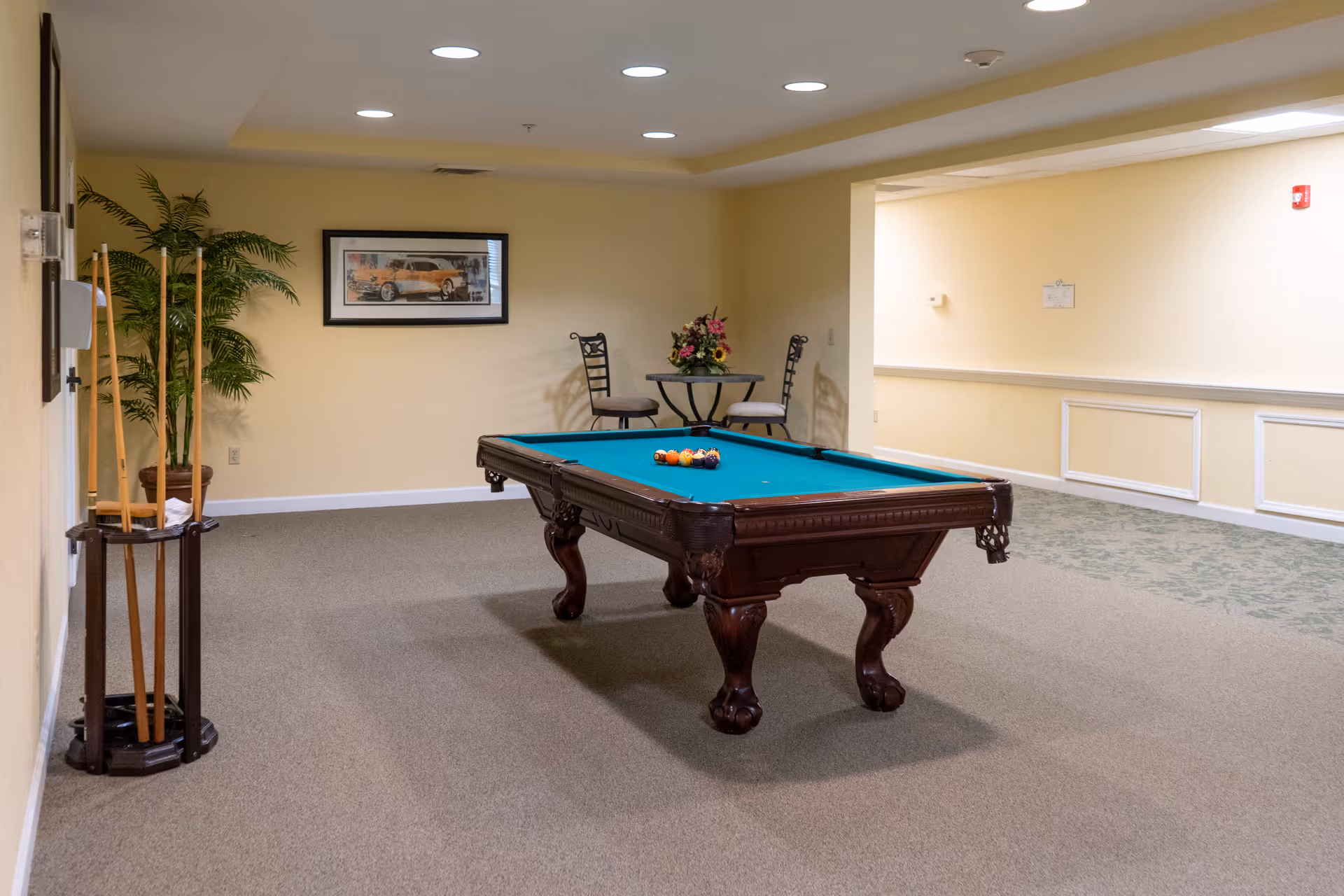 Indoor recreational room with a pool table in the center, a rack holding pool cues on the left, a small round table with two chairs and a flower arrangement in the back corner, beige walls, carpeted floor, and a framed picture on the wall.