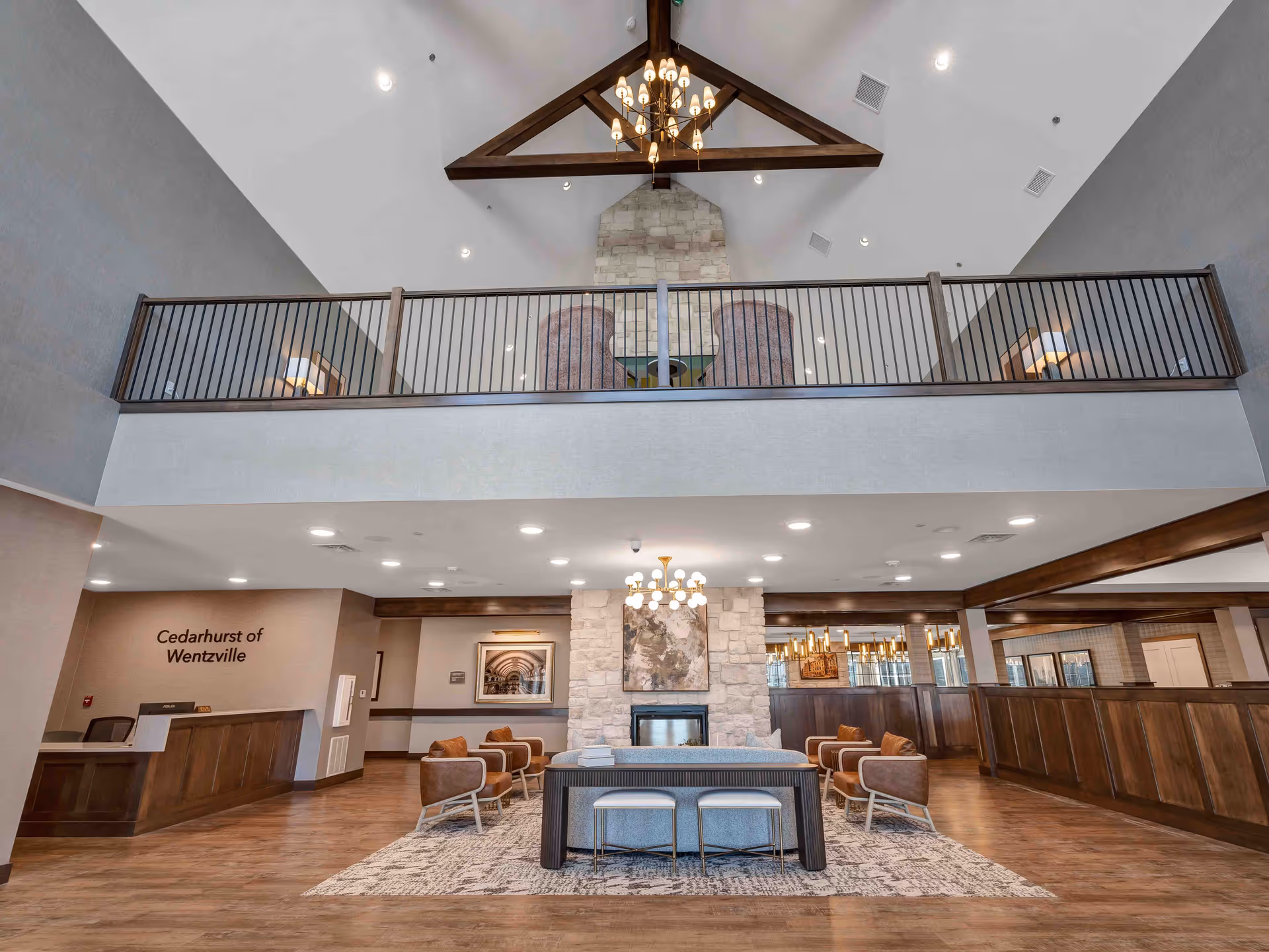 Spacious senior living facility lobby with high vaulted ceiling featuring wooden beams and a chandelier. The room includes a stone fireplace with artwork above it, comfortable seating with brown chairs and a gray sofa, a patterned rug, and a reception desk on the left with the text 'Cedarhurst of Wentzville' on the wall. There is a balcony area above with additional seating and lamps.