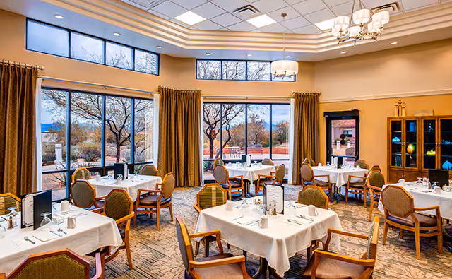 A bright dining room with multiple tables covered in white tablecloths, set with glasses, napkins, and menus. The room has large windows with views of trees and outdoor scenery, beige walls, and a chandelier hanging from the ceiling. There are green cushioned chairs around the tables and a wooden cabinet with glass doors in the corner.
