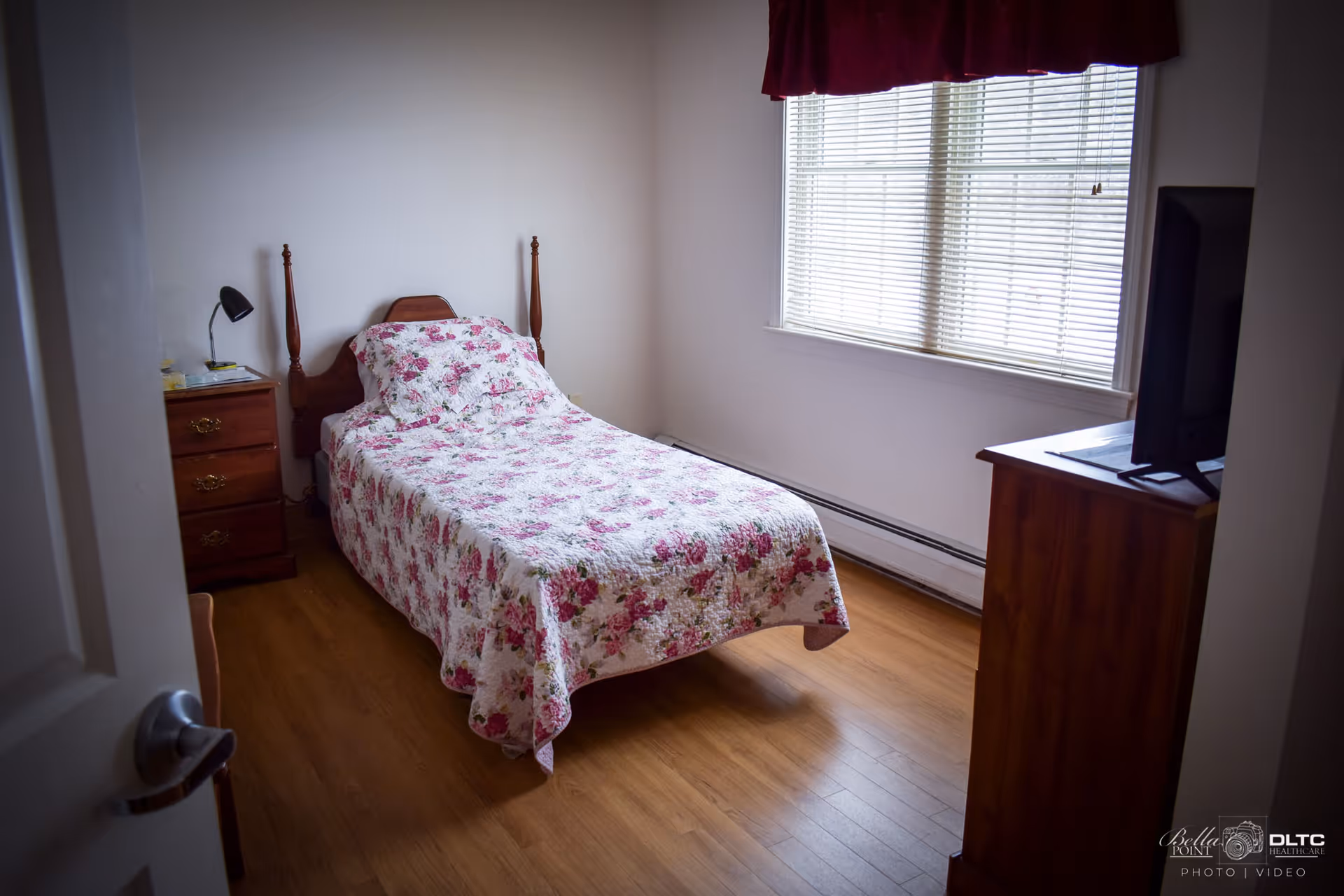 A small bedroom with a single bed covered in a floral quilt. There is a wooden nightstand with a lamp and some items on it next to the bed. A wooden dresser with a flat-screen TV on top is positioned near a window with closed blinds and a dark red valance. The floor is wooden, and the walls are plain white.