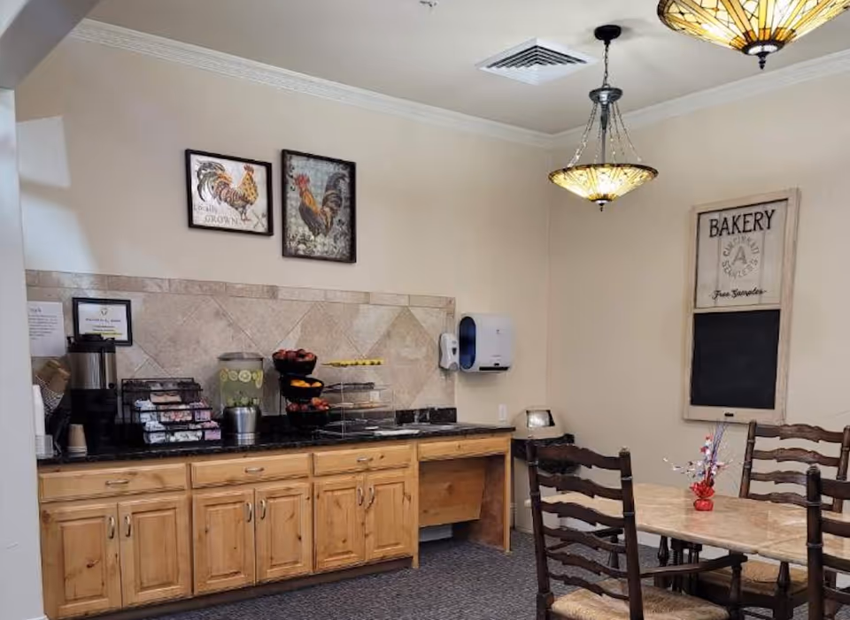 Small dining area with wooden cabinets and a countertop stocked with beverages and snacks, a table with chairs, wall art and pendant lights.