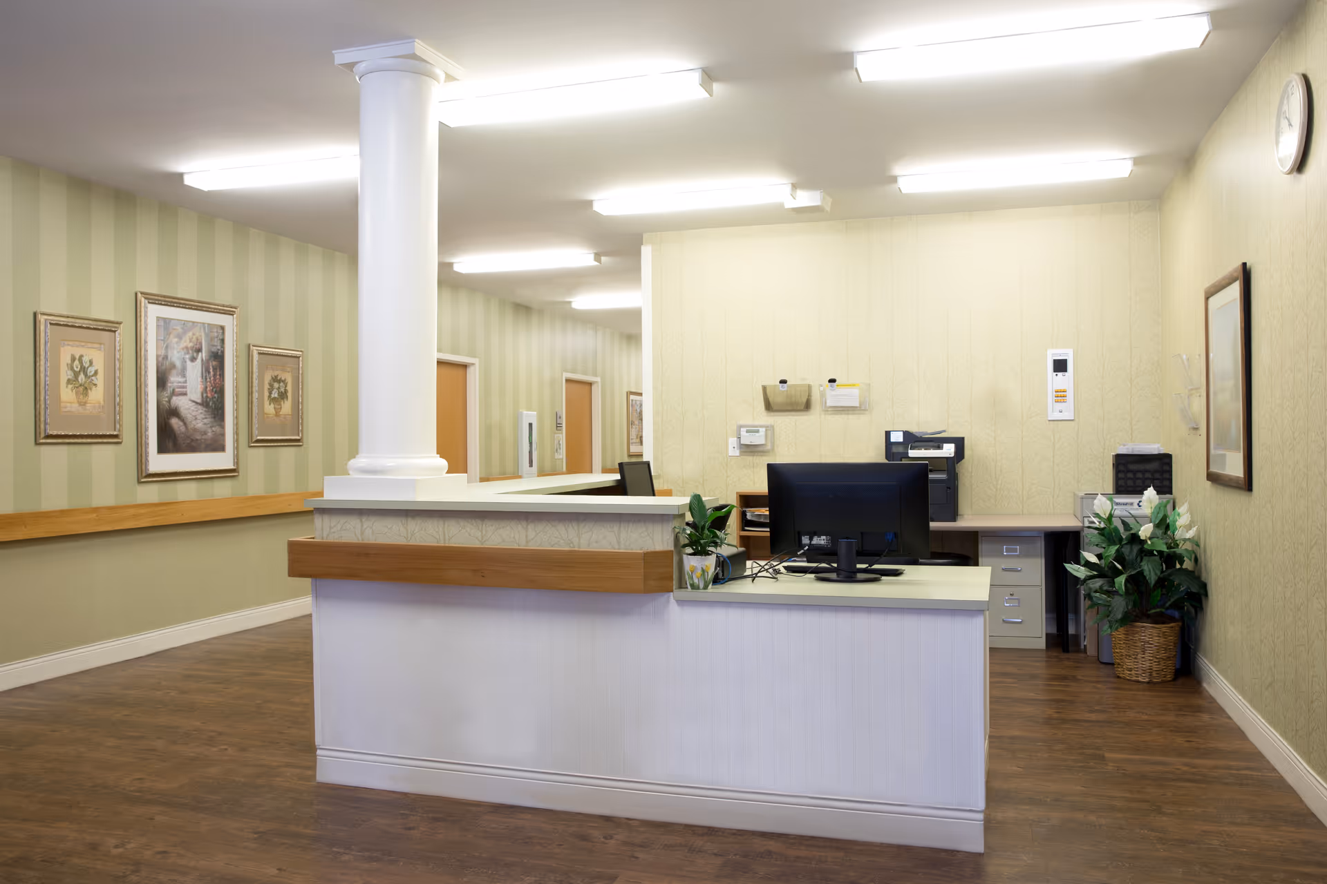 Reception desk and office area with a computer monitor, column, framed artwork, and potted plants in a senior living facility.