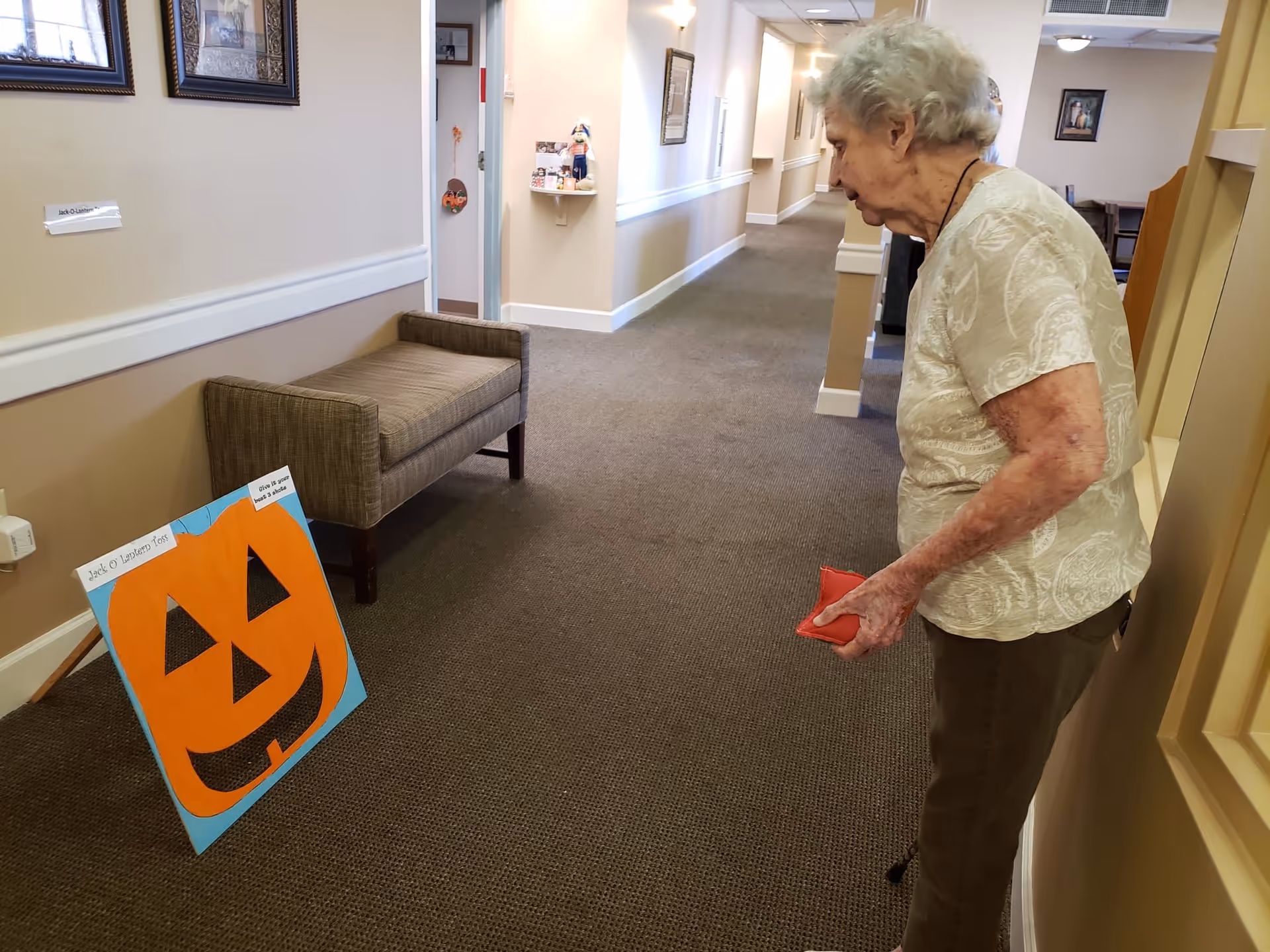 An elderly woman stands in a hallway of a senior living facility, looking at a large orange paper pumpkin decoration labeled 'Jack O Lantern Toss' placed on the floor. The hallway has beige walls, carpeted floor, framed pictures on the walls, and a bench against one wall.
