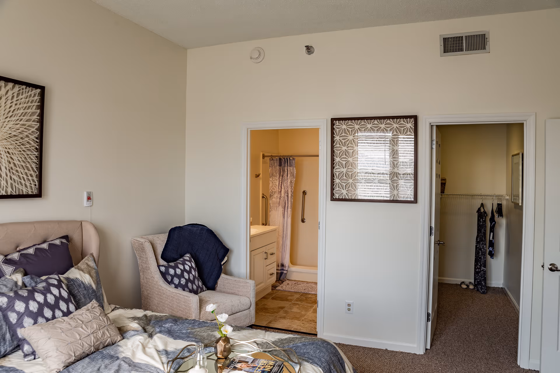 A bedroom in a senior living facility with a bed covered in patterned pillows and blankets, a beige armchair with a dark throw blanket, and two doorways leading to a bathroom with a shower curtain and a walk-in closet with hanging clothes.