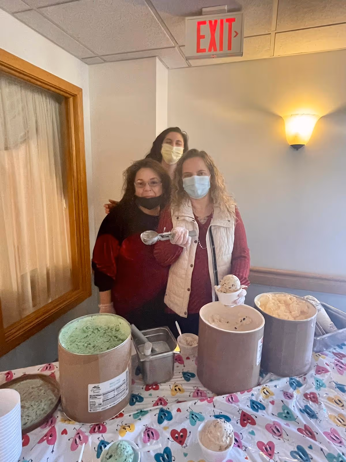 Three women wearing face masks and gloves standing behind a table with large containers of ice cream and scoops, inside a room with a window and an exit sign above the door.