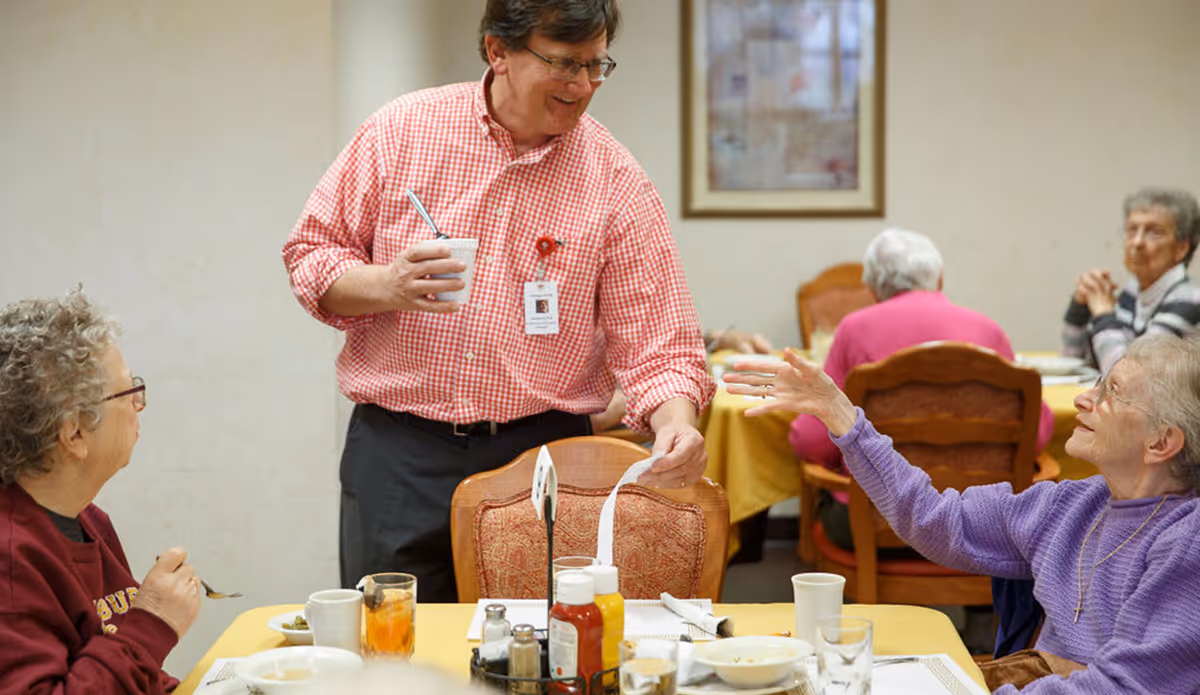 A staff member in a red checked shirt interacts with elderly residents seated around a dining table in a communal dining room.
