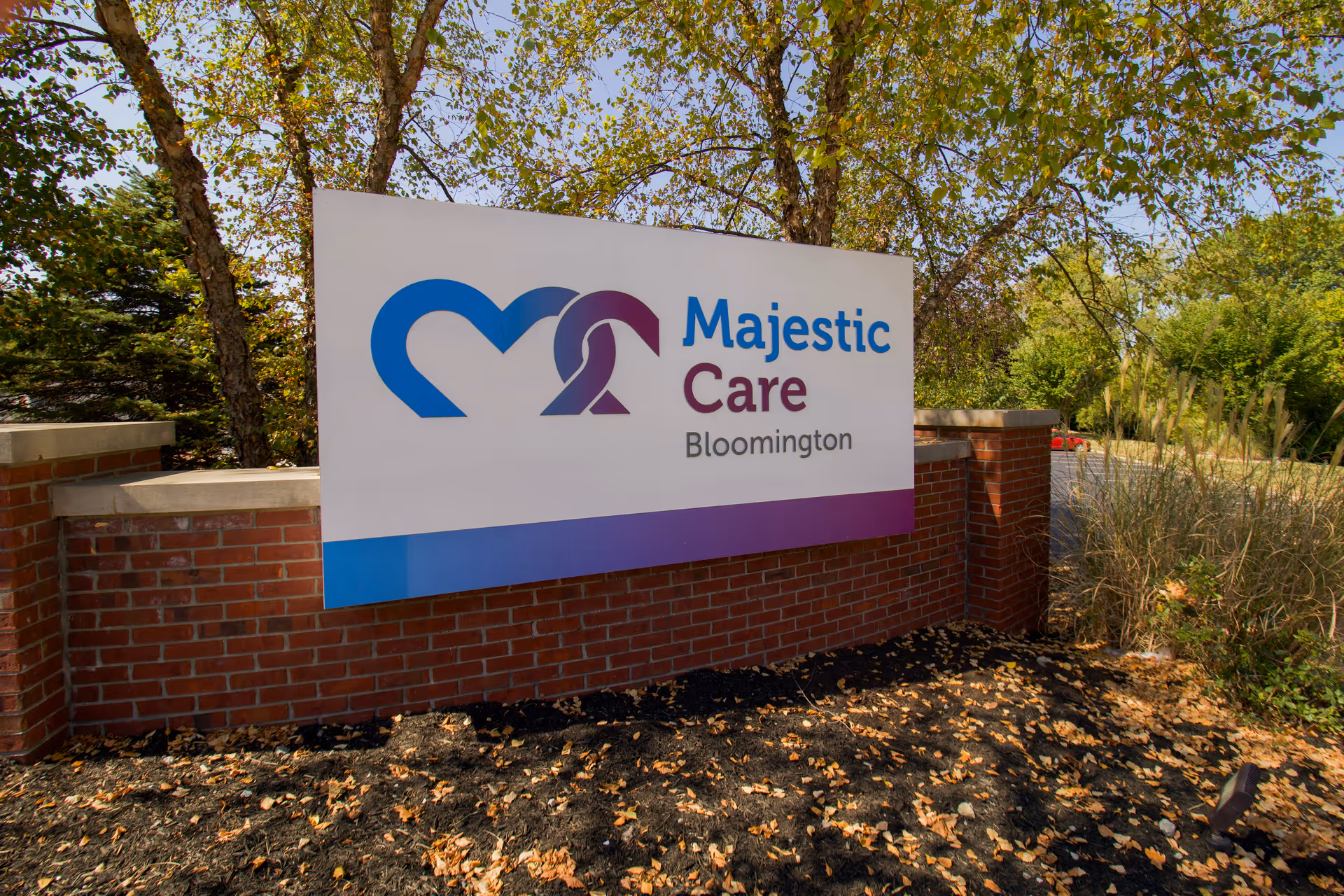 Outdoor sign for Majestic Care Bloomington mounted on a brick base with trees and greenery in the background.
