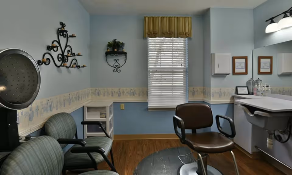 A small room with light blue walls and wood flooring, featuring a window with white blinds and a yellow valance. There are three chairs, including a salon-style chair in front of a white sink with a large mirror above it. The walls have decorative wallpaper border and metal wall shelves with small plants and candle holders.