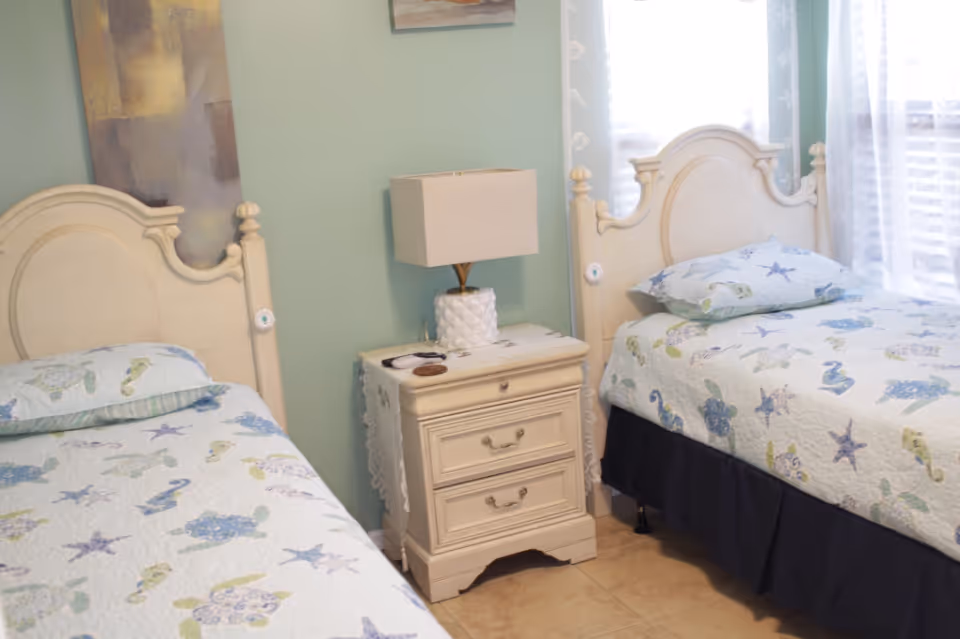 Two twin beds with sea-themed quilts separated by a nightstand and lamp in a softly painted bedroom.