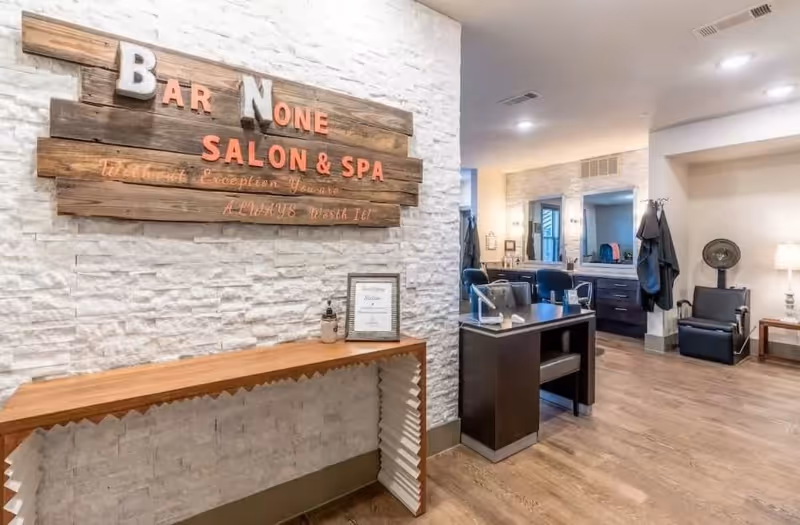 Interior view of a salon and spa area with a wooden sign on a white textured wall that reads 'Bar None Salon & Spa'. The room features a wooden console table beneath the sign, a manicure station with chairs, mirrors, and salon equipment. The floor is wood, and the space is well-lit with ceiling lights and a table lamp.