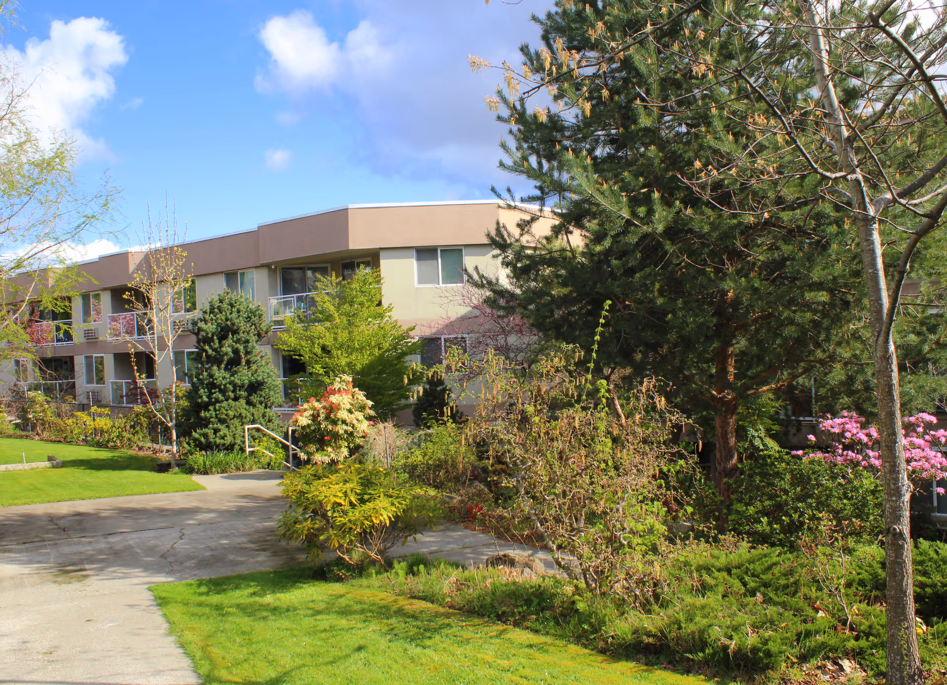 Exterior view of a two-story retirement living facility named Boulevard Park Place Active Retirement Living, surrounded by lush greenery, trees, and shrubs under a partly cloudy blue sky.