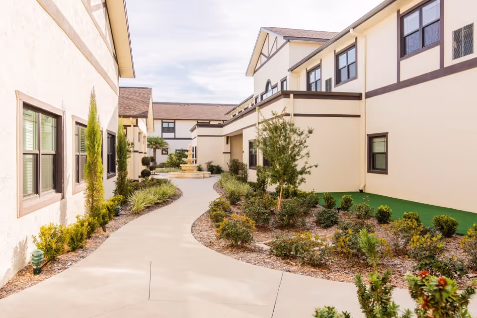 Outdoor walkway at Royal Dalton House with beige buildings on both sides, landscaped garden beds with shrubs and small trees, and a fountain visible in the distance under a partly cloudy sky.