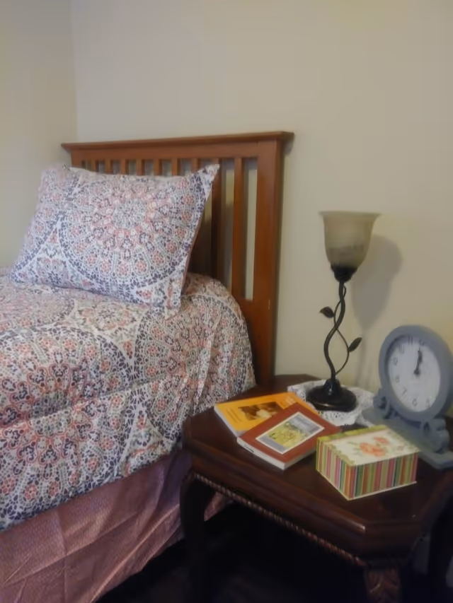 A wooden bed with a patterned bedspread and matching pillow next to a wooden nightstand. The nightstand holds a decorative lamp, a round clock, and several books and a small striped box.