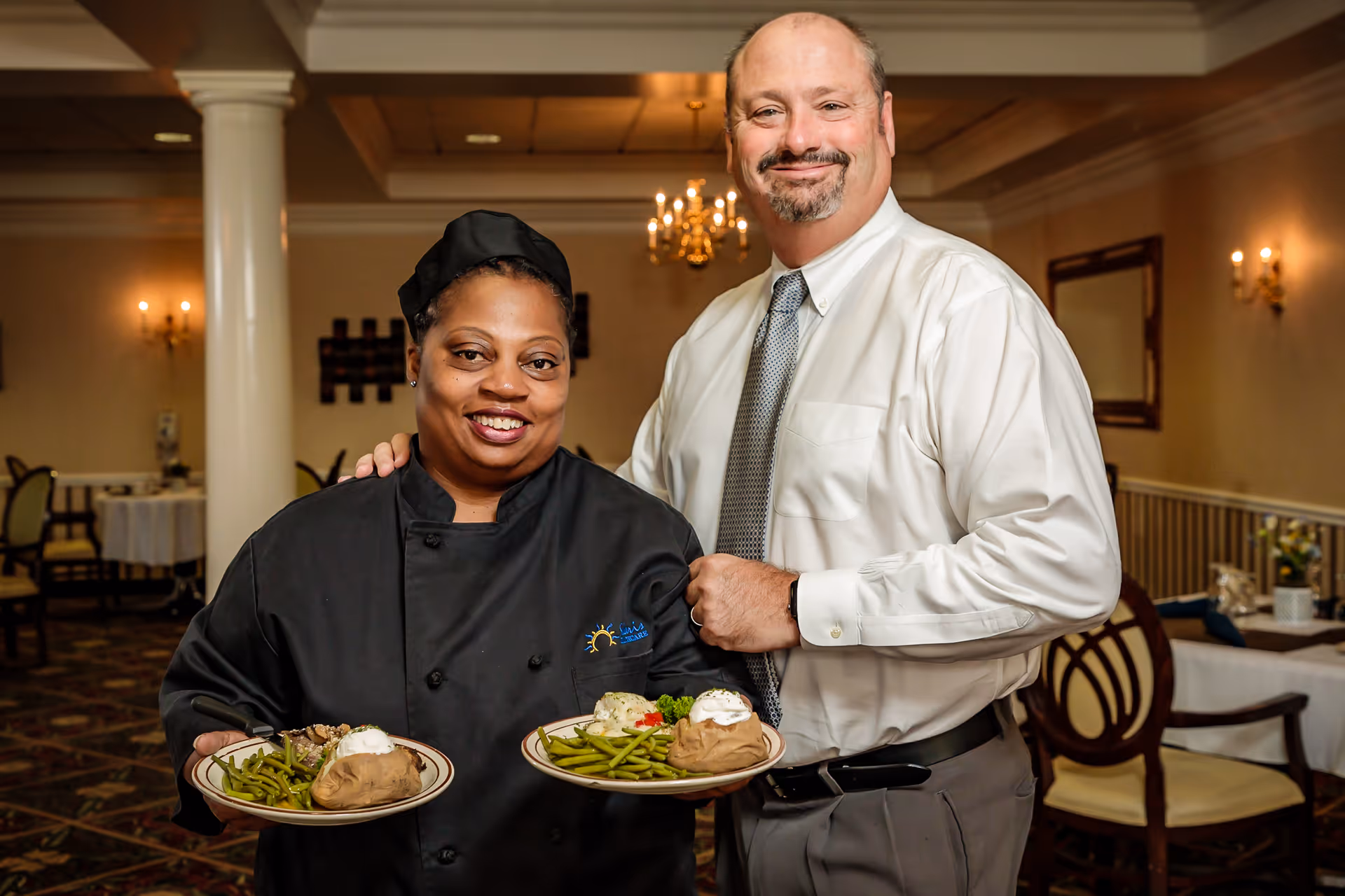 A smiling female chef in a black uniform and a man in a white shirt and tie stand together in a dining room. The chef holds two plates of food, each with green beans, a baked potato topped with sour cream, and a serving of meat. The dining room has elegant decor with chandeliers, wall sconces, and tables set for meals.