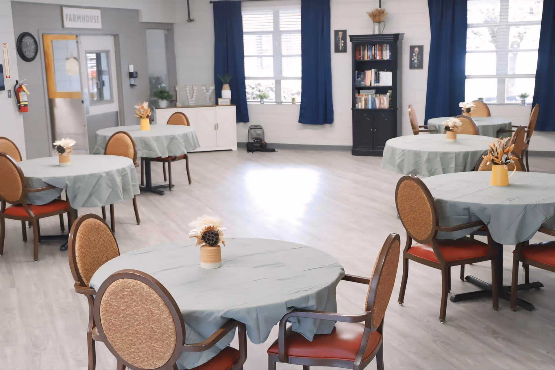 A bright dining room with several round tables covered with light green tablecloths, each table decorated with a small vase of dried flowers. The room has wooden chairs with cushioned seats and backs, light wood flooring, large windows with blue curtains, a black bookshelf filled with books, and a white cabinet. A sign above a door reads 'FARMHOUSE'.