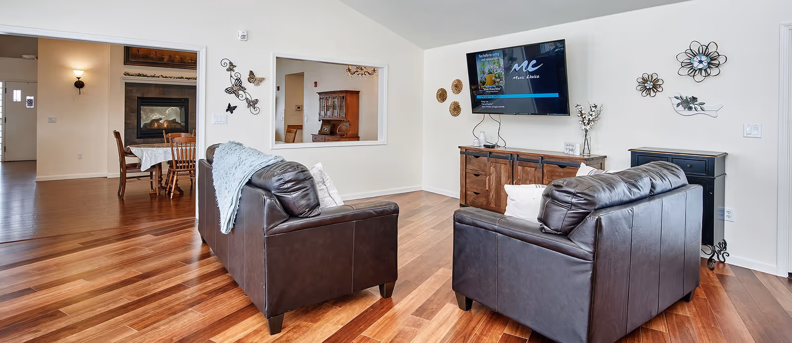 A senior living facility common area with two dark brown leather sofas facing a wall-mounted flat screen TV. The room has wooden flooring and white walls decorated with metal floral wall art. In the background, there is a dining area with a wooden dining table and chairs, and a fireplace.
