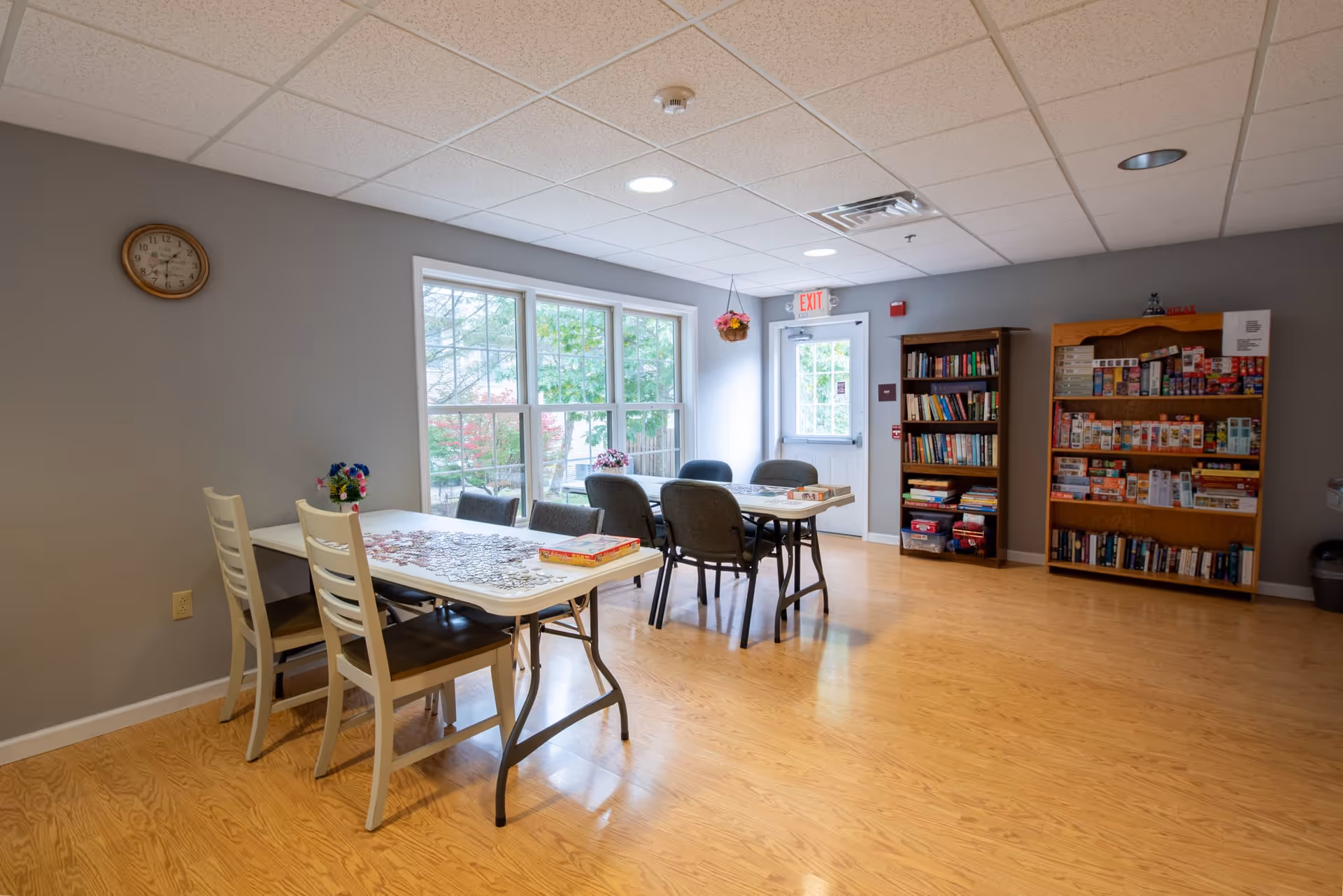 A bright activity room with two tables and several chairs. One table has a partially completed jigsaw puzzle and a puzzle box on it. The room has large windows letting in natural light, a clock on the wall, and two bookshelves filled with books and board games. There is a door with an exit sign above it and a hanging flower basket near the door.