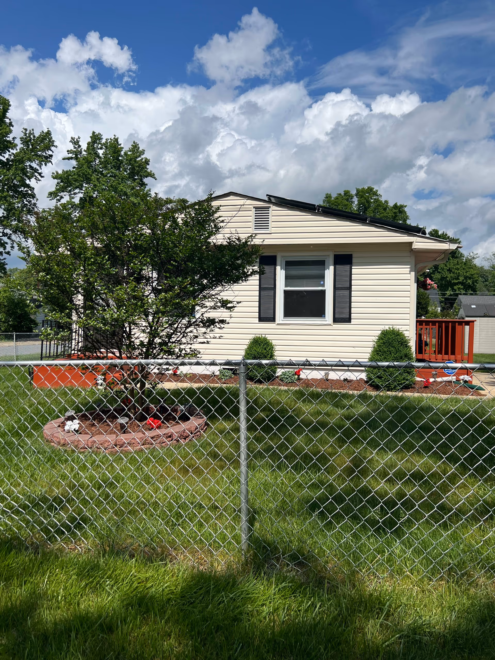 A small beige house with black shutters and a single window, surrounded by a green lawn and a chain-link fence. There is a tree and some small bushes in front of the house, with a partly cloudy blue sky in the background.