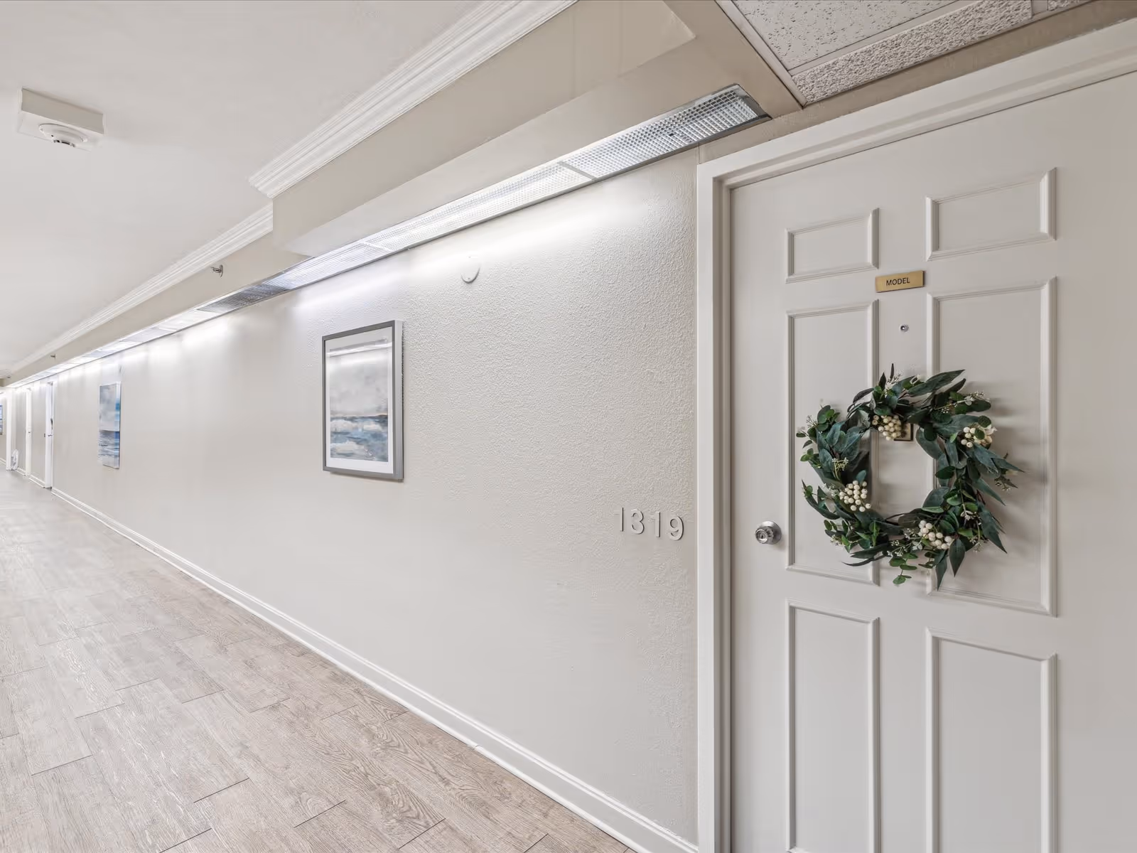 Bright interior hallway showing a white apartment door with a decorative wreath and framed artwork on the wall.