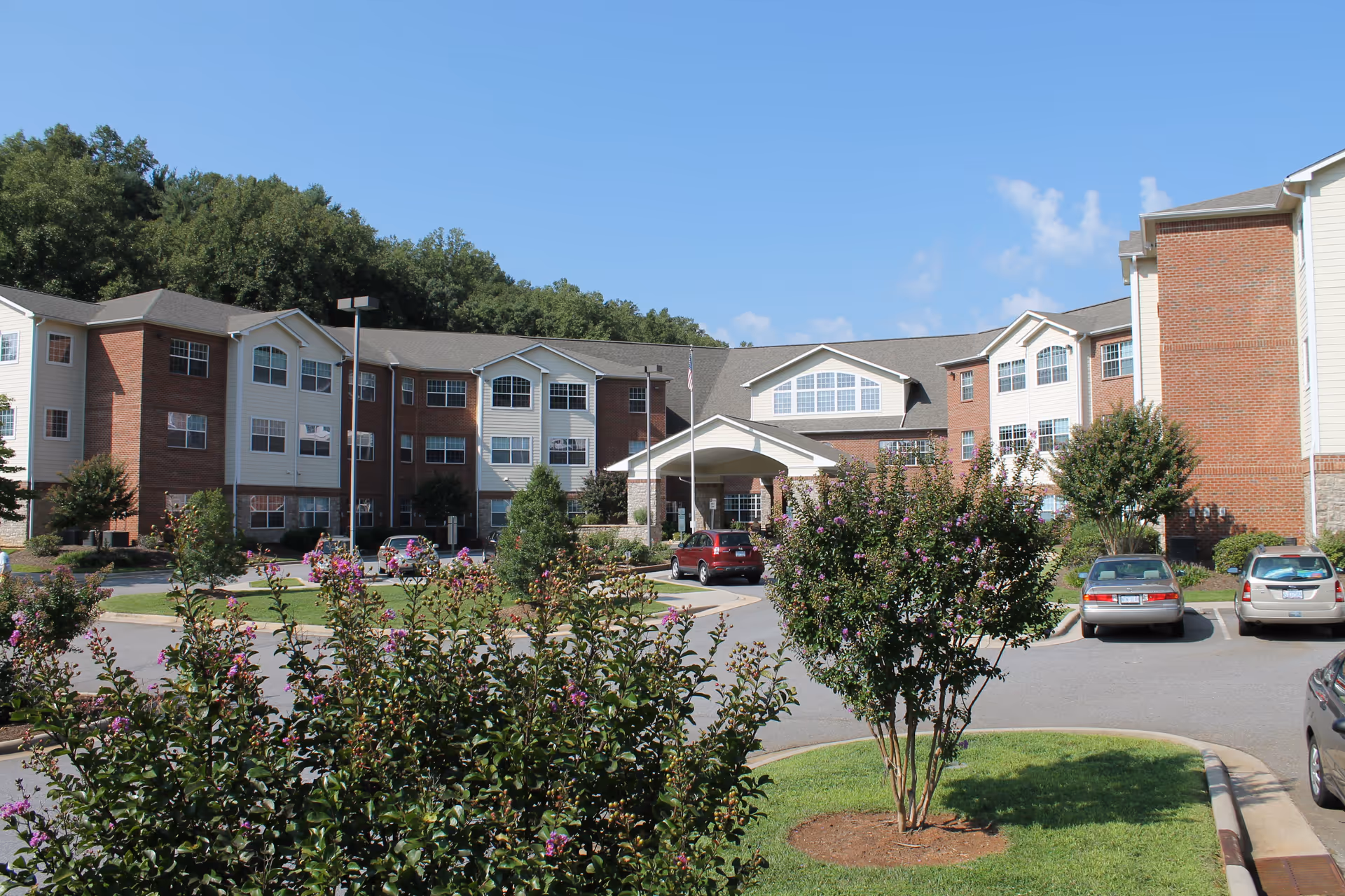 Exterior view of a multi-story senior living facility named Rose Glen Village with a parking lot in front, surrounded by green trees and flowering bushes under a clear blue sky.