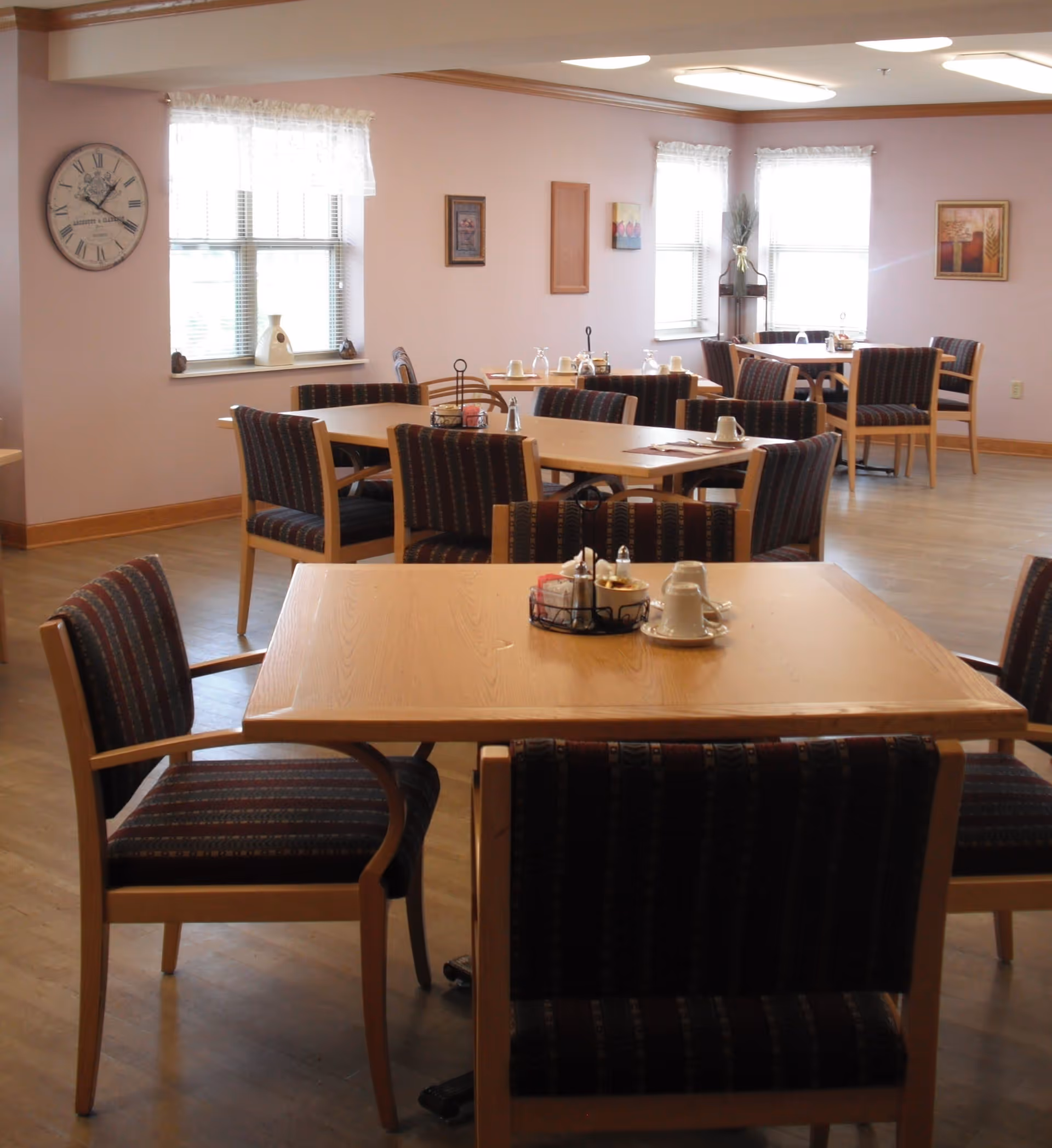 Communal dining room with wooden tables and striped upholstered chairs, place settings, windows, and a wall clock.