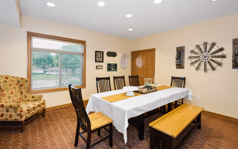 A dining room with a rectangular table covered with a white tablecloth and a yellow runner. The table is surrounded by six wooden chairs and a wooden bench. On the table are decorative items including a tray, a glass container, and a cake stand. The room has a patterned carpet, beige walls with various wall decorations including a large metal windmill piece, and a window with blinds showing an outdoor view. There is also a patterned armchair near the window.