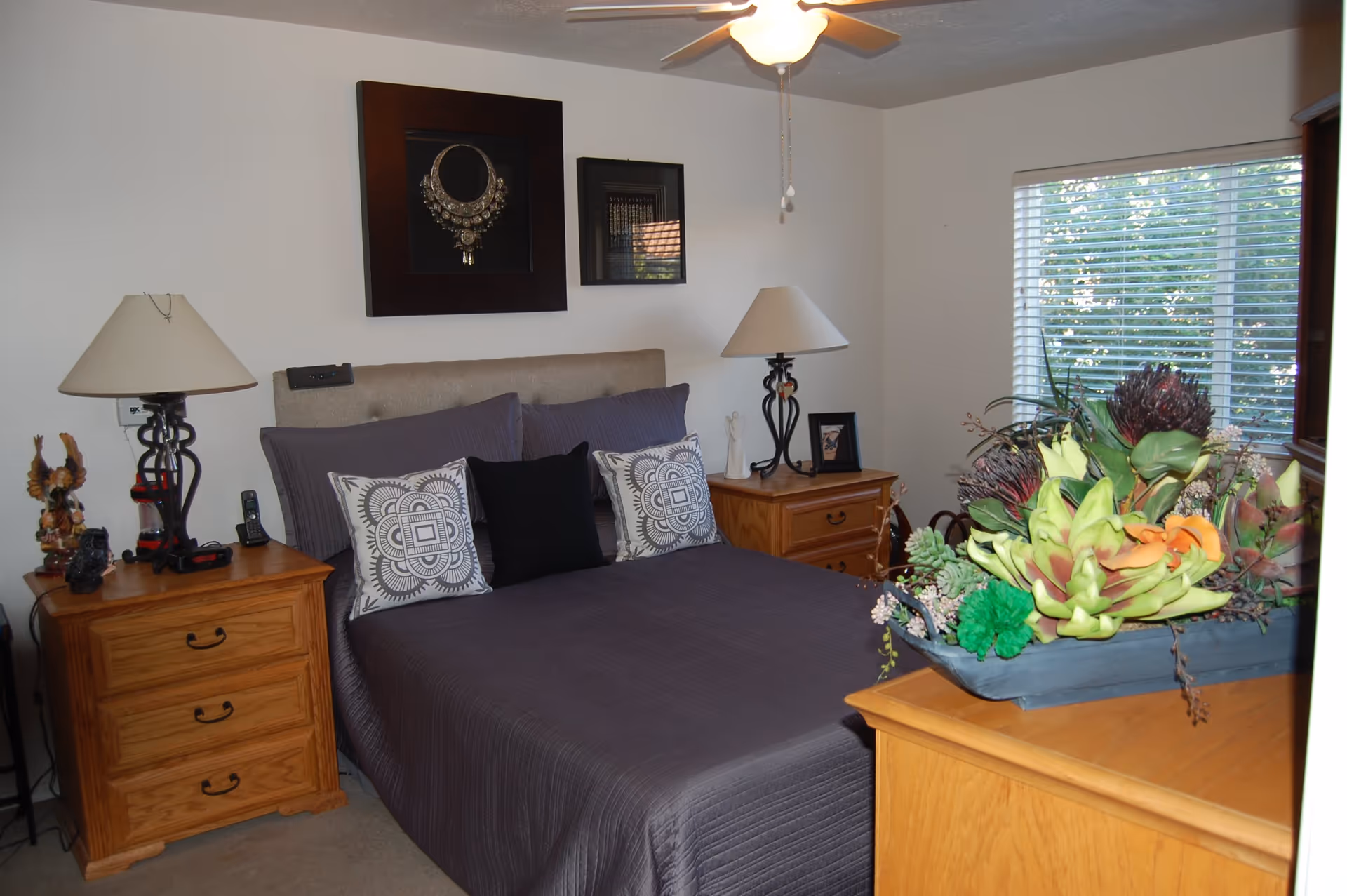 A cozy bedroom with a neatly made bed covered in a dark gray quilt and decorative pillows. There are two wooden nightstands on either side of the bed, each with a lamp and various decorative items. Above the bed are two framed pieces of wall art. A window with blinds lets in natural light, and a wooden dresser with a large floral arrangement is visible in the foreground.