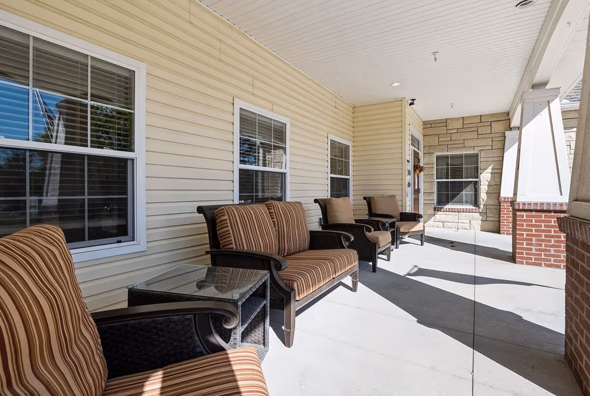 Covered outdoor porch area with cushioned chairs and a loveseat arranged along the wall of a building with beige siding and windows. The porch has a concrete floor and white ceiling with recessed lighting, and brick and stone accents on the supporting columns and walls.
