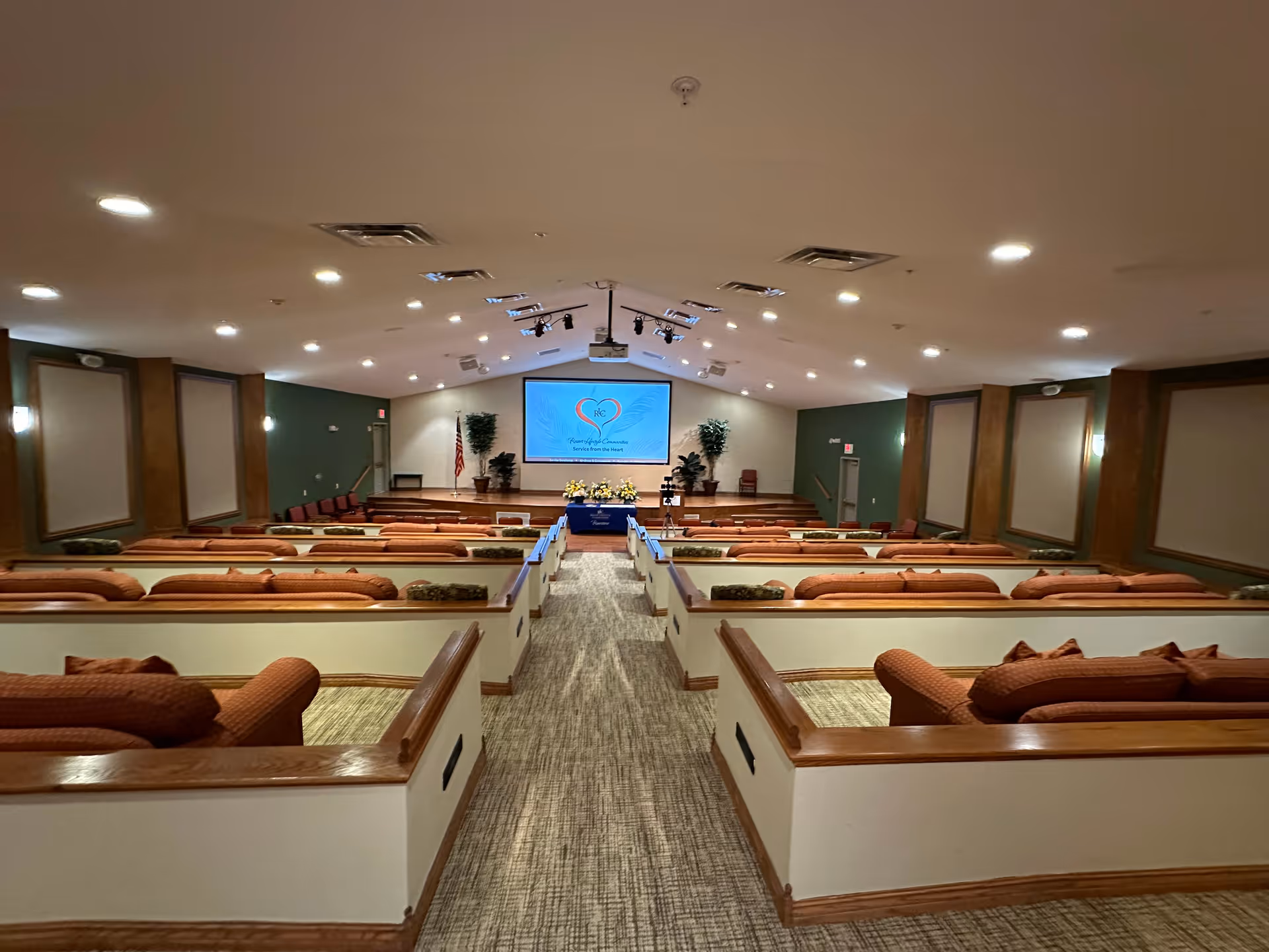A large interior auditorium with tiered cushioned bench seating facing a stage and projection screen at the front.