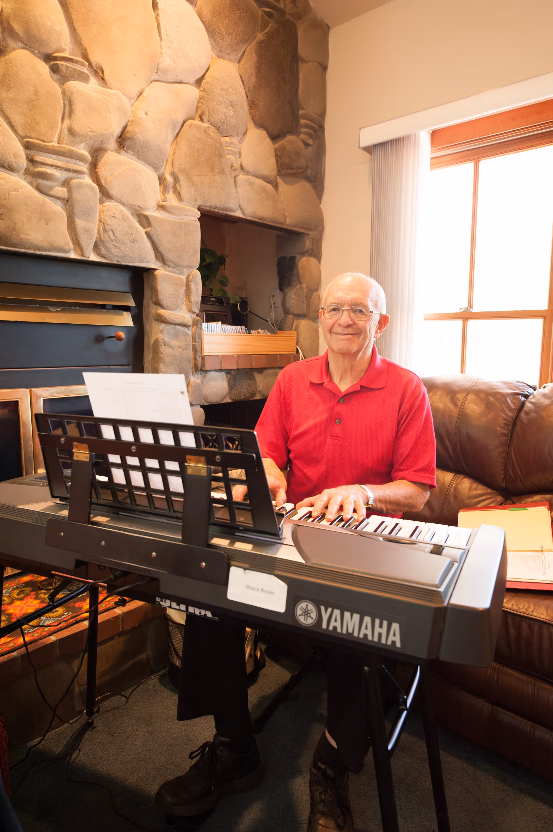 An elderly man wearing a red polo shirt is sitting and playing a Yamaha keyboard in a cozy living room with a stone fireplace and a large window with vertical blinds. There is a brown leather couch beside him and some papers on it.