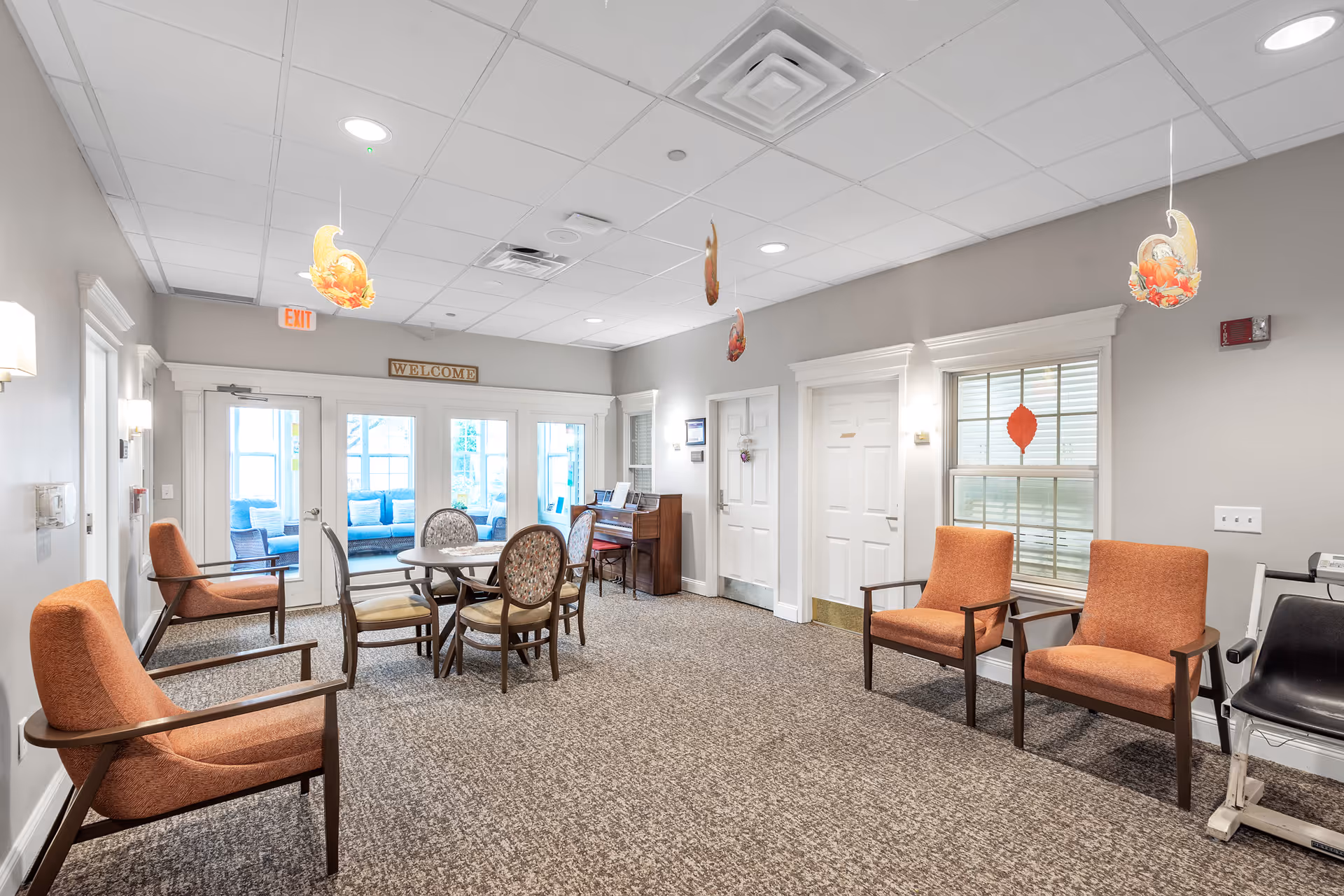 Well-lit senior living common room with orange armchairs, a round table and chairs, a piano, and glass doors under a 'WELCOME' sign.