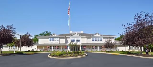 Front exterior view of Shawnee Estates Senior Living building with a circular driveway, landscaped garden in the center, and two flags on a flagpole against a clear blue sky.