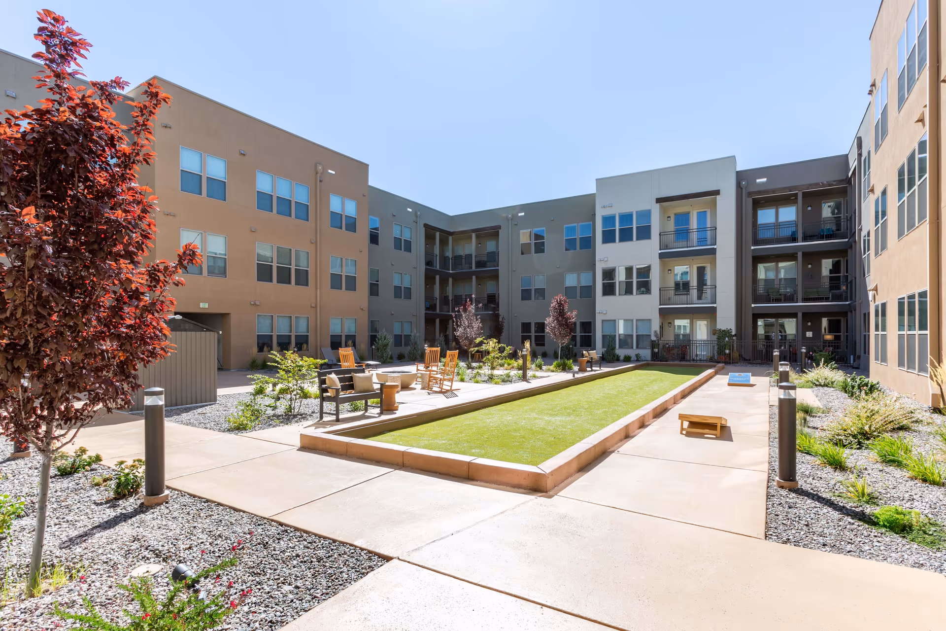 Outdoor courtyard area of a senior living facility with a bocce ball court in the center, surrounded by benches, rocking chairs, small tables, and landscaped plants and trees. The courtyard is enclosed by a three-story building with multiple windows and balconies under a clear blue sky.
