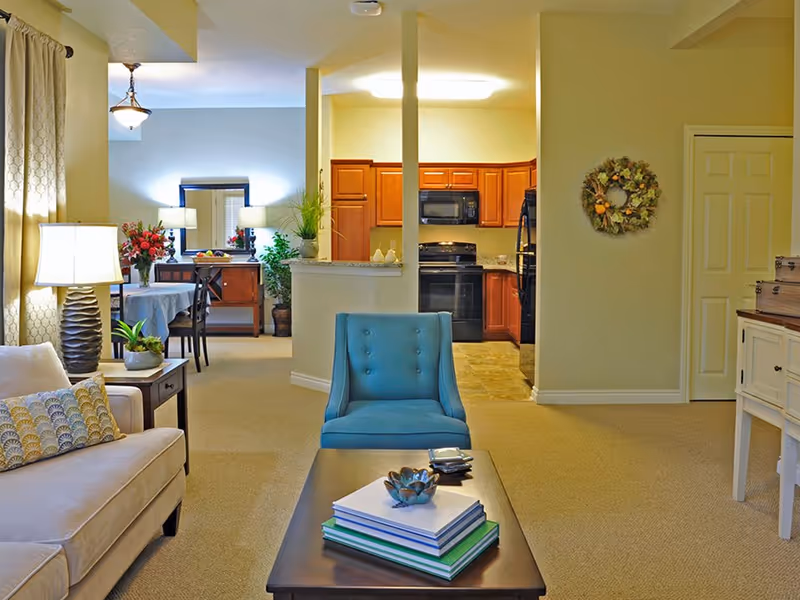 A cozy senior living interior featuring a beige sofa with patterned pillows, a blue armchair, and a dark wooden coffee table with books and decorative items. In the background, there is a dining area with a table, chairs, and a sideboard with lamps and plants. The kitchen area with wooden cabinets, a black stove, microwave, and refrigerator is visible beyond a half wall with two vertical columns. The walls are painted light yellow, and there is a decorative wreath hanging on one wall.