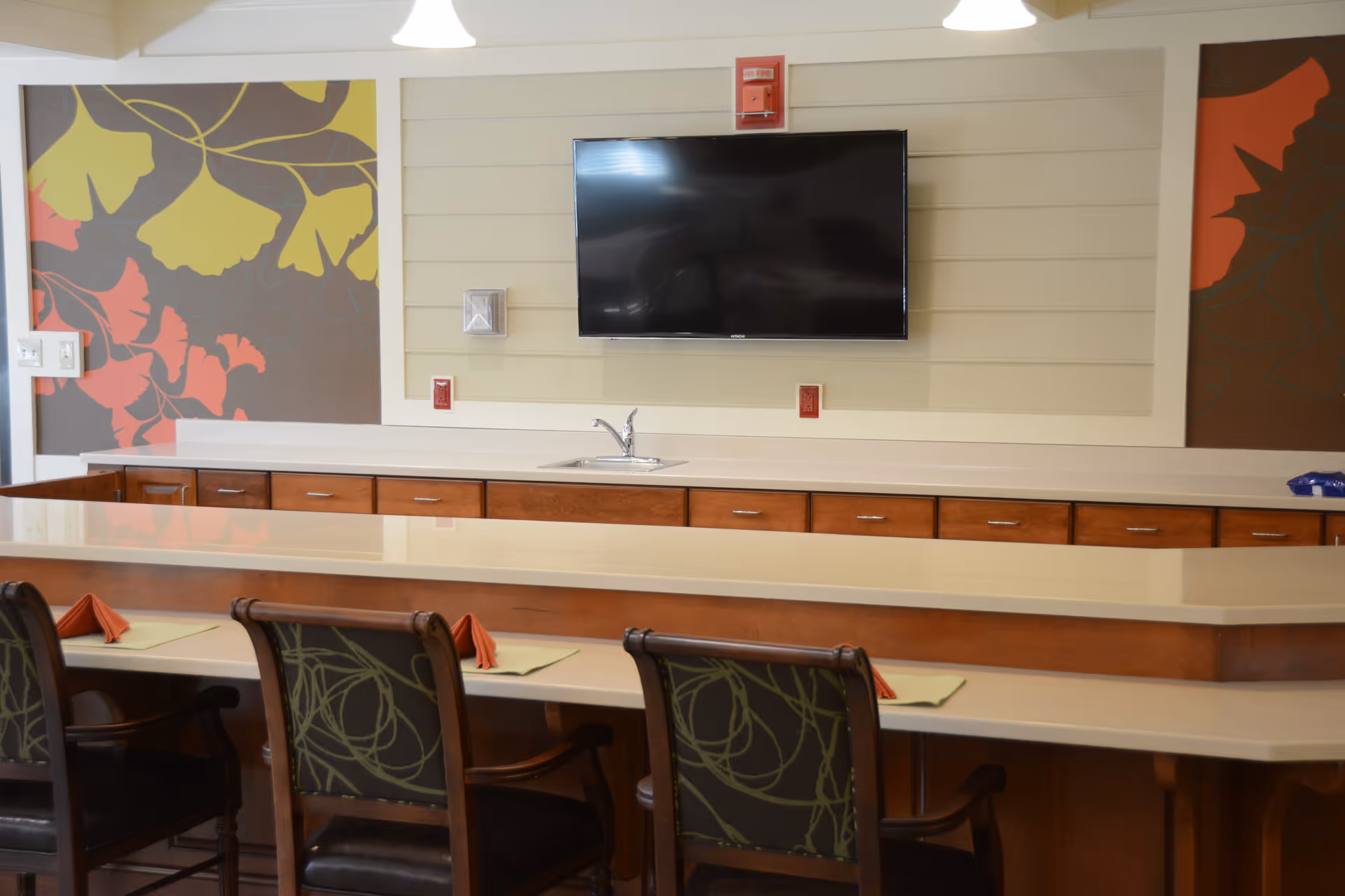 Long communal dining counter with chairs, placemats, a sink and a wall-mounted TV against decorative wall panels.