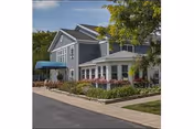 Exterior view of a two-story building with gray siding and white trim, surrounded by greenery and flowers. There is a blue awning over the entrance and a paved driveway in front.