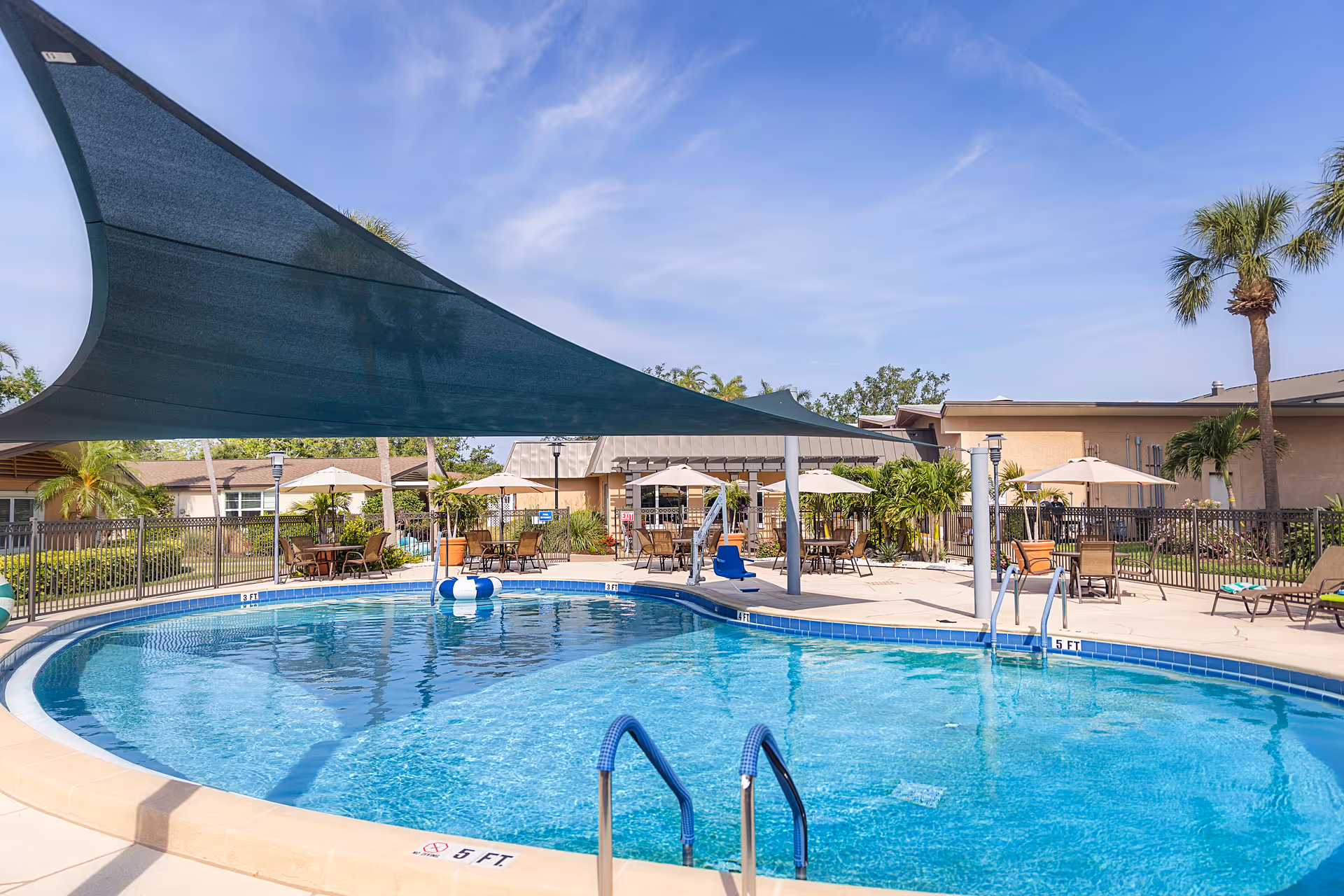 Outdoor swimming pool area with blue water, surrounded by a concrete deck with tables, chairs, umbrellas, and palm trees under a clear blue sky.
