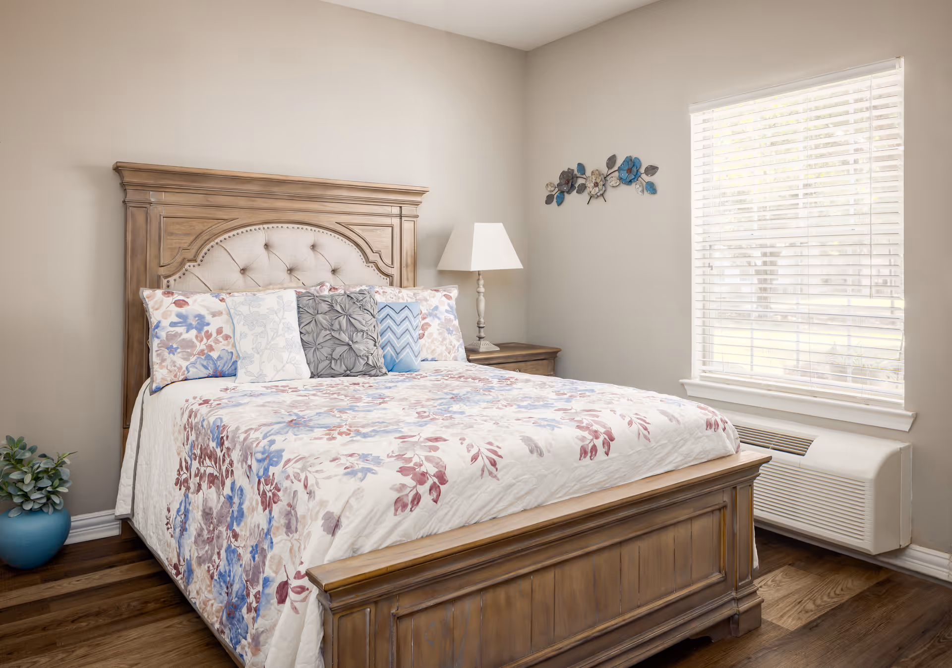 A bright bedroom with a wooden bedframe, floral bedding, nightstand with a lamp, and a window with blinds.