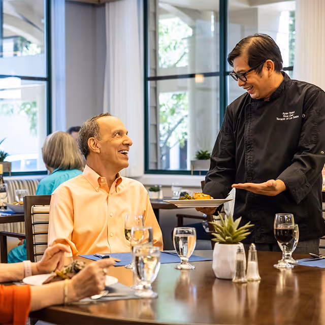 A smiling waiter in a black uniform serving a plate of food to a happy elderly man seated at a dining table in a bright, modern dining room with large windows and other diners in the background.