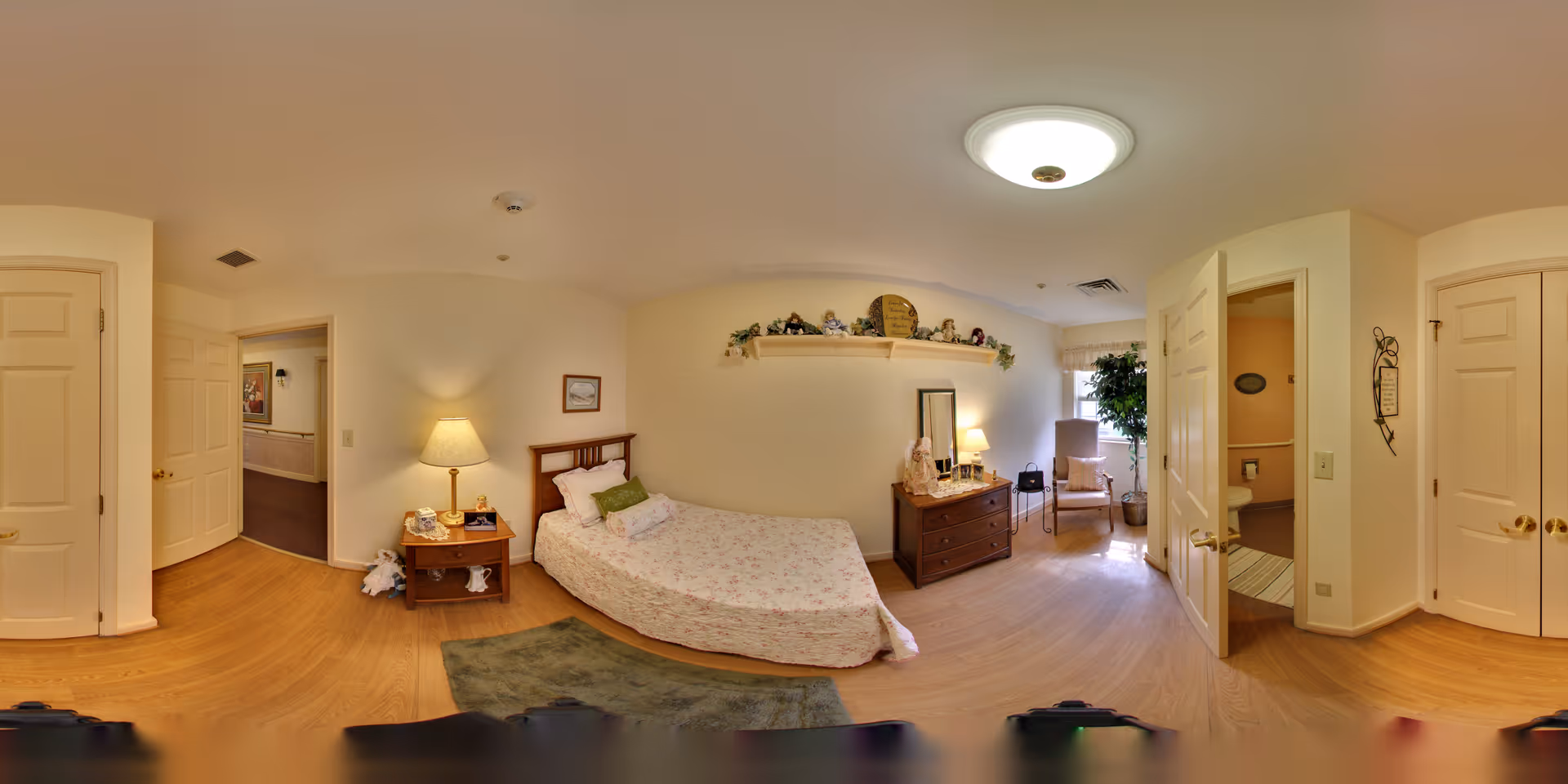 A cozy senior living bedroom with a single bed covered in a floral bedspread, a wooden nightstand with a lamp and phone, a wooden dresser with a mirror and decorative items, a green rug on the wooden floor, and a chair near a window with a potted plant. The room has cream-colored walls and multiple doors leading to other areas.
