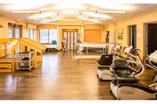 A spacious rehabilitation room with exercise equipment including stationary bikes and parallel bars, large windows letting in natural light, and a wooden floor. The room is well-lit with ceiling lights and has a warm, inviting atmosphere.