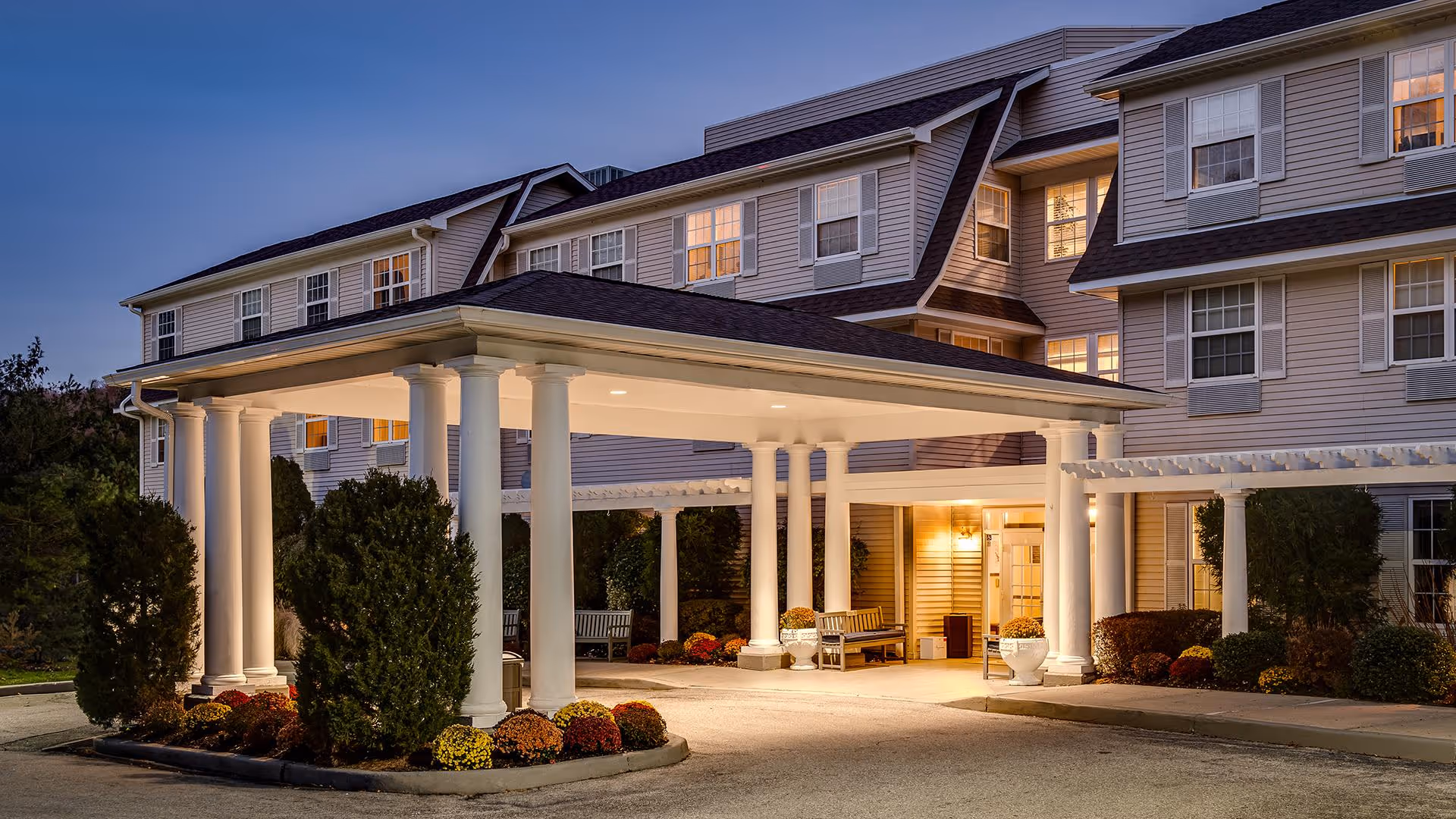 Front entrance of a multi-story senior living building with a covered porte-cochere, white columns, benches, and landscaping at dusk.