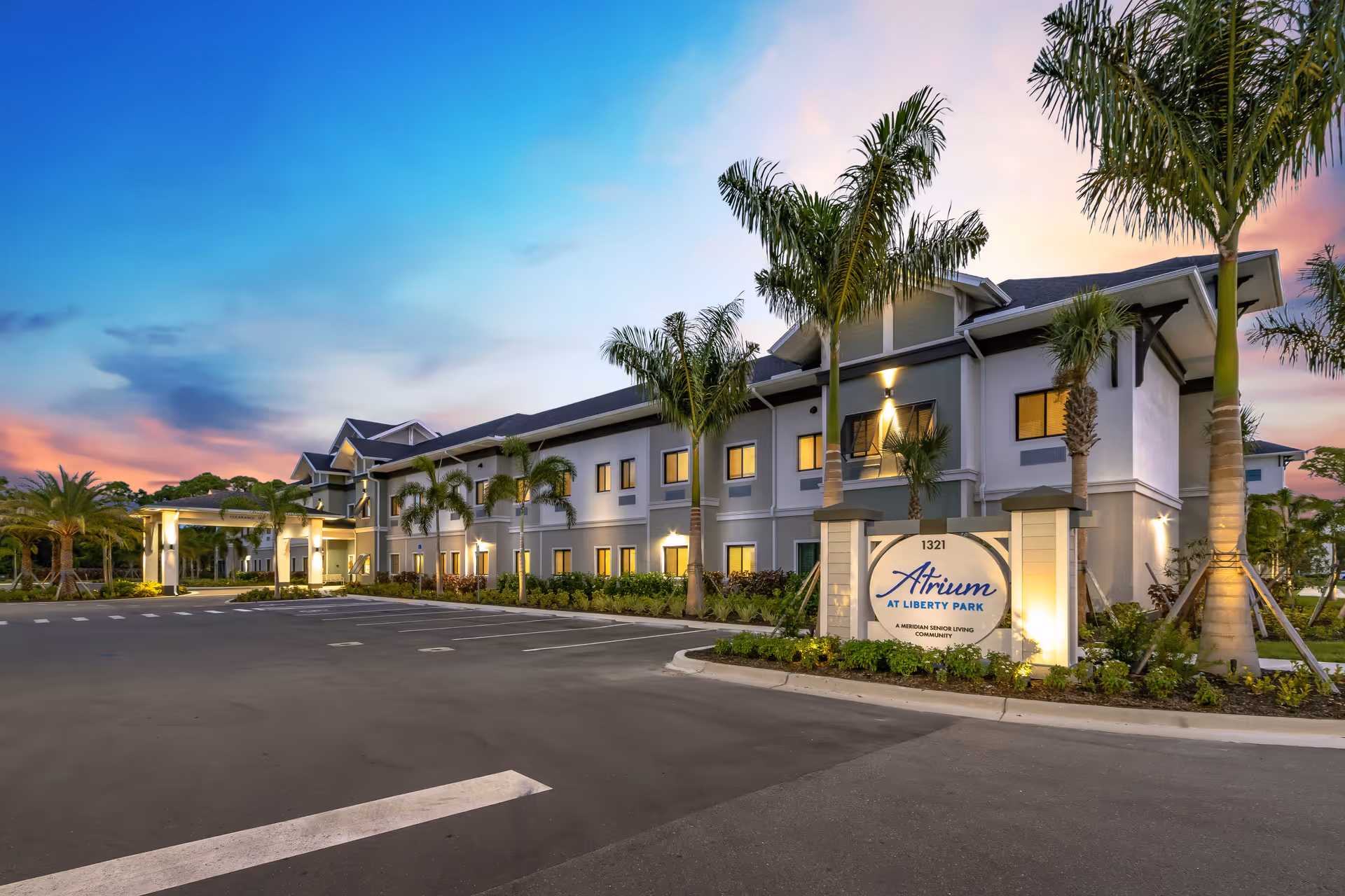 Exterior view of Atrium at Liberty Park senior living facility at dusk, showing a two-story building with lit windows, palm trees, a parking lot, and a sign with the facility name.