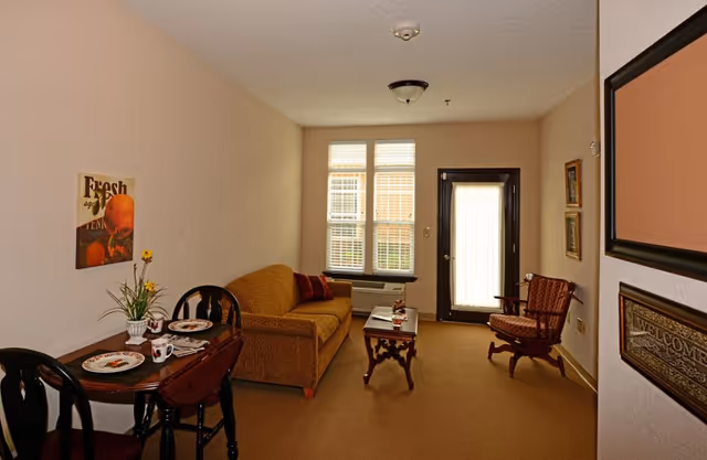 Small furnished living room with a sofa, armchair, coffee table, and a small dining table by a window and door.