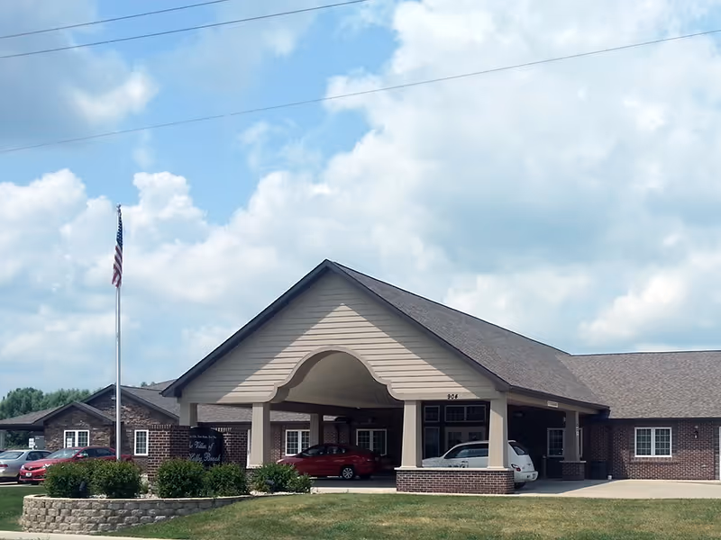 Exterior view of Villas of Holly Brook Effingham building with a covered entrance, several parked cars, an American flag on a flagpole, and a partly cloudy sky.